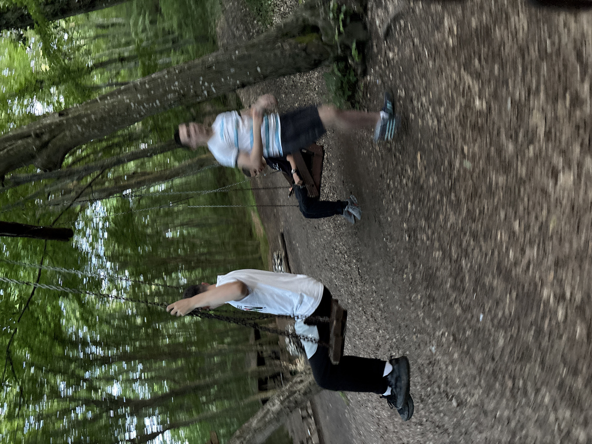 The image depicts a group of individuals on swings in a forest. The perspective is low and wide-angle, creating a distorted effect, especially on the ground.
In the foreground, on the left, a person is seated on a wooden swing, their body blurred by motion. They are wearing a white t-shirt, black pants, and black sneakers with white socks. Their left hand is visible, gripping the swing's chain.
Further back and to the right, another wooden swing is occupied by two individuals. One, in the foreground of this swing, is wearing a black t-shirt and black pants, and appears to be holding a phone. The other, behind them, is visible only as a blur.
In the background, a third individual is standing, blurred by motion, wearing a white t-shirt with blue stripes and dark shorts. They are positioned to the right of the swings.
The environment is a dense forest with tall trees and lush green foliage creating a canopy overhead. The ground is covered with dirt, leaves, and small stones, also distorted by the wide-angle lens and motion. Some wooden structures are faintly visible in the background, suggesting a park or recreational area within the forest.
The overall impression is one of dynamic movement and outdoor activity in a natural setting. There is no specific indication of the city of Kichevo, Bulgaria, in the image itself.