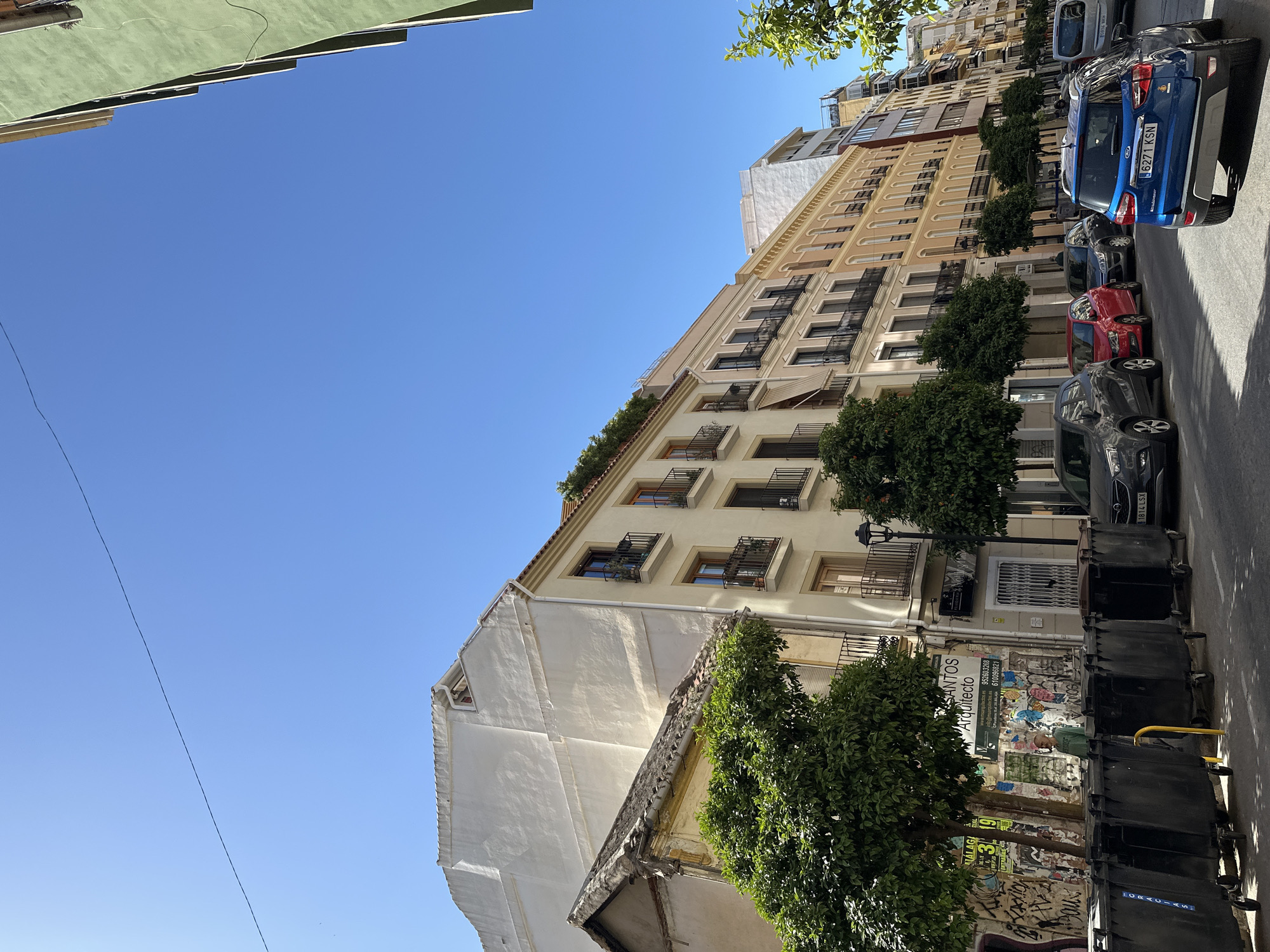 The image depicts a street in Málaga, Spain. The view is angled upwards, showcasing several buildings with cream-colored facades and dark window frames. Some buildings have small balconies with wrought iron railings. Green trees line the street, partially obscuring the buildings. Parked cars of various colors (blue, red, grey) are visible on the right side of the street. Black trash bins line the sidewalk. Graffiti covers a wall between the trees. The sky is a clear blue.