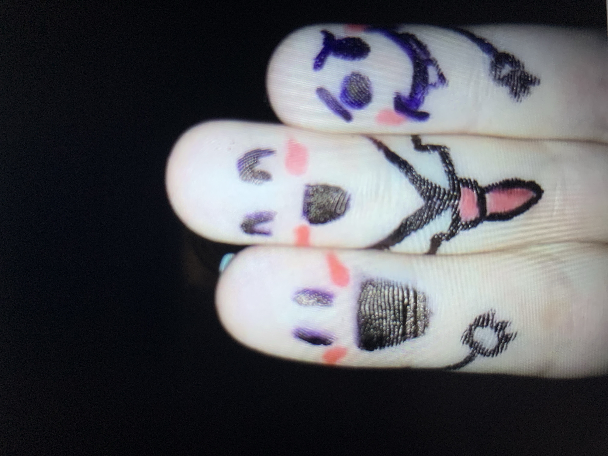 Three fingers have been decorated with faces drawn using markers. The finger on the left has purple eyes, pink circles on its cheeks, and a black, textured mouth. The middle finger has wide, purple eyes with black pupils, pink blush marks on its cheeks, a wide black smile, and is wearing a white shirt with a black tie that has pink detailing. The finger on the right has purple, lopsided eyes, a purple wavy mouth, and pink circles on its cheeks. The background is black. The image does not provide any information about the city of Lymanka, Ukraine.