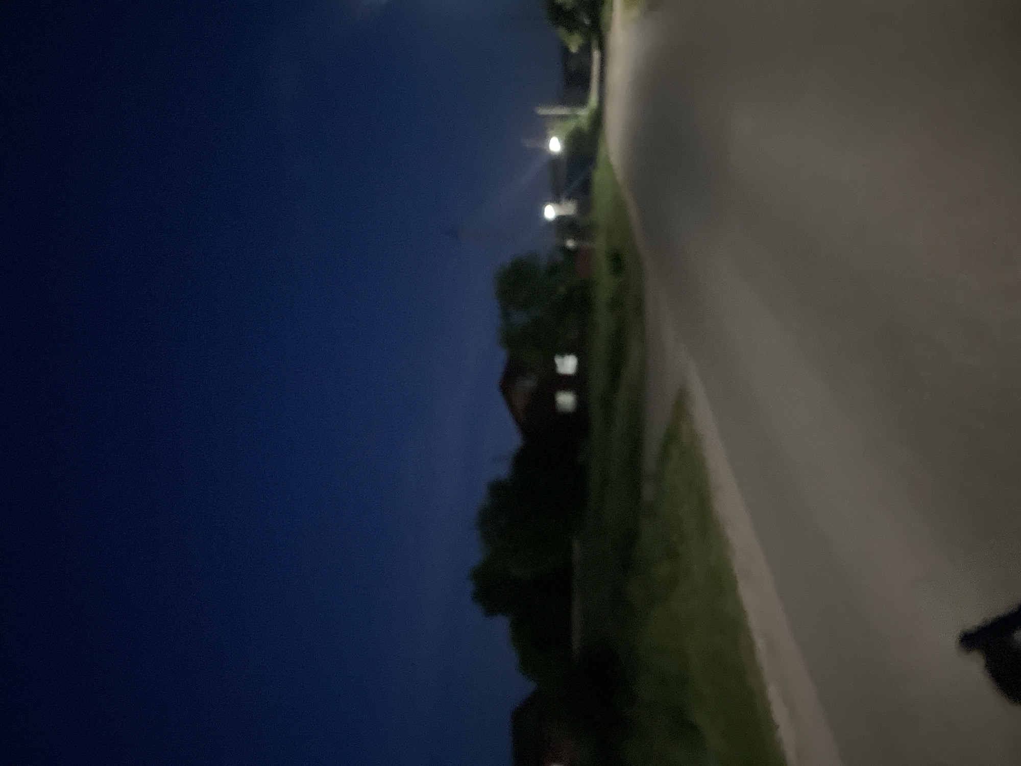 The image depicts a road in Красные Баки, Russia, at dusk. The sky is a deep blue, suggesting the transition from daylight to night. On the left side of the road, there is a grassy embankment and some greenery. In the background, several houses are visible, some with illuminated windows, indicating habitation. Trees are scattered throughout the scene, casting dark shadows. Two streetlights are illuminated, casting cones of light onto the road. The road surface appears to be asphalt and is relatively smooth. In the foreground, a dark, blurry silhouette of what appears to be a person on a bicycle is visible on the road, suggesting movement. The overall atmosphere is calm and quiet, typical of a residential area at the end of the day.