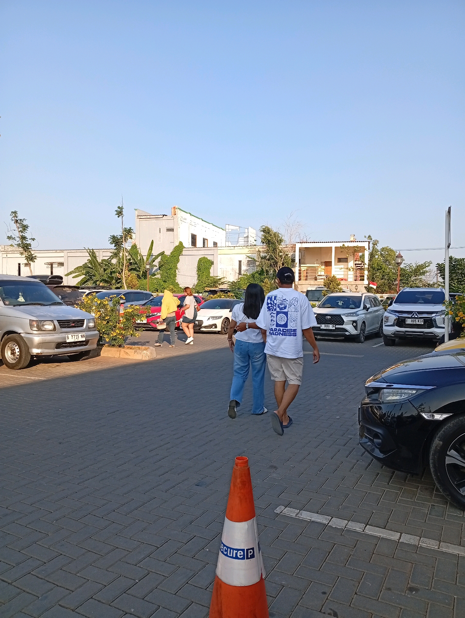 A man and a woman walk away from the camera in a paved parking lot in Indramayu, Indonesia, with several cars parked around them. The man wears a white t-shirt with blue graphics, tan shorts, and blue flip-flops. The woman wears a white t-shirt, blue jeans, and white shoes. They are walking towards a white SUV and a white and silver SUV. Behind them, a silver van is parked on the left. In the background, a white building with a covered patio is visible, surrounded by trees and foliage. The sky is clear and blue. An orange traffic cone with blue and white lettering that reads "secure P" is in the foreground.