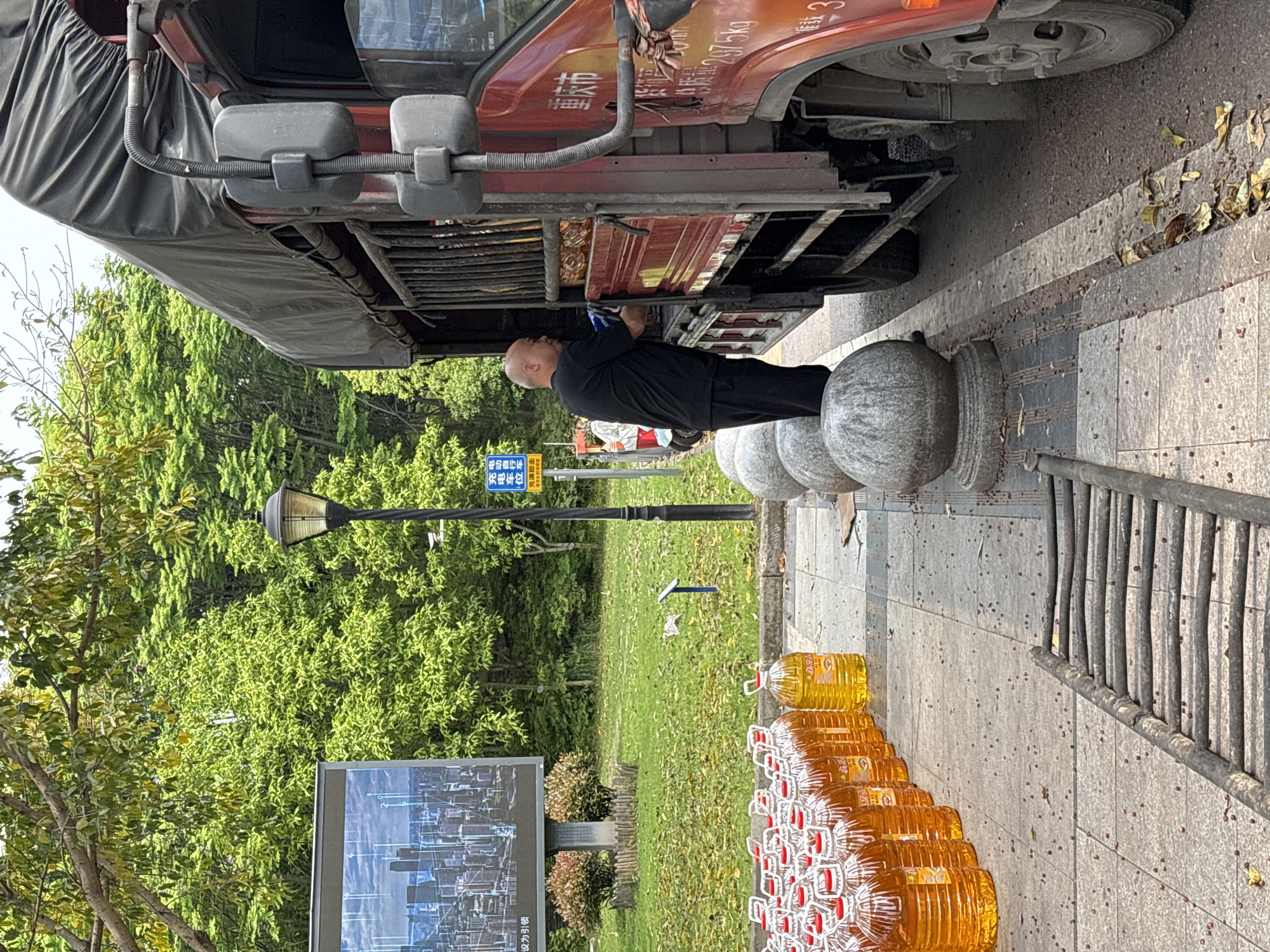 The image depicts a street scene in Jiangjin, China. A bald man in black clothing is standing beside a red truck, appearing to work on its underside. The truck is parked on a sidewalk alongside a row of round concrete bollards and a metal fence. Next to the fence is a long row of large, clear plastic bottles filled with a yellow liquid, possibly oil or another liquid product. In the background is a grassy area with a street lamp and an advertising billboard. A street sign is visible in the distance. The ground is paved and there are scattered leaves and debris.