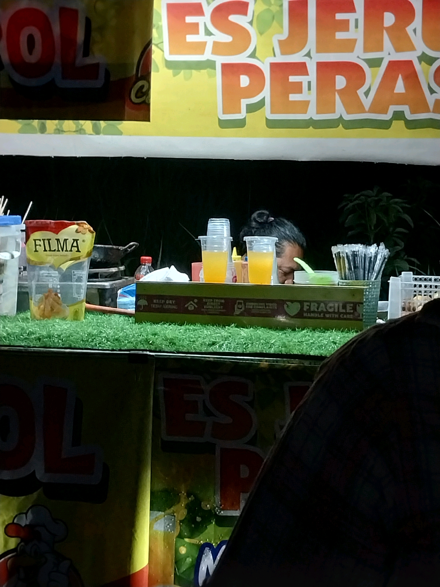 The image depicts a food stall at night, likely in a bustling area of Nglegok, Indonesia. A large, brightly colored banner with the words "ES JERUK PERAS" (Freshly Squeezed Orange Juice) is prominently displayed. The stall is set up with various items, including cups of orange juice, a packet of "FILMA" brand oil, and a small frying pan on a stove. A person, partially visible, appears to be working at the stall. The overall scene suggests a casual, outdoor food vending operation.