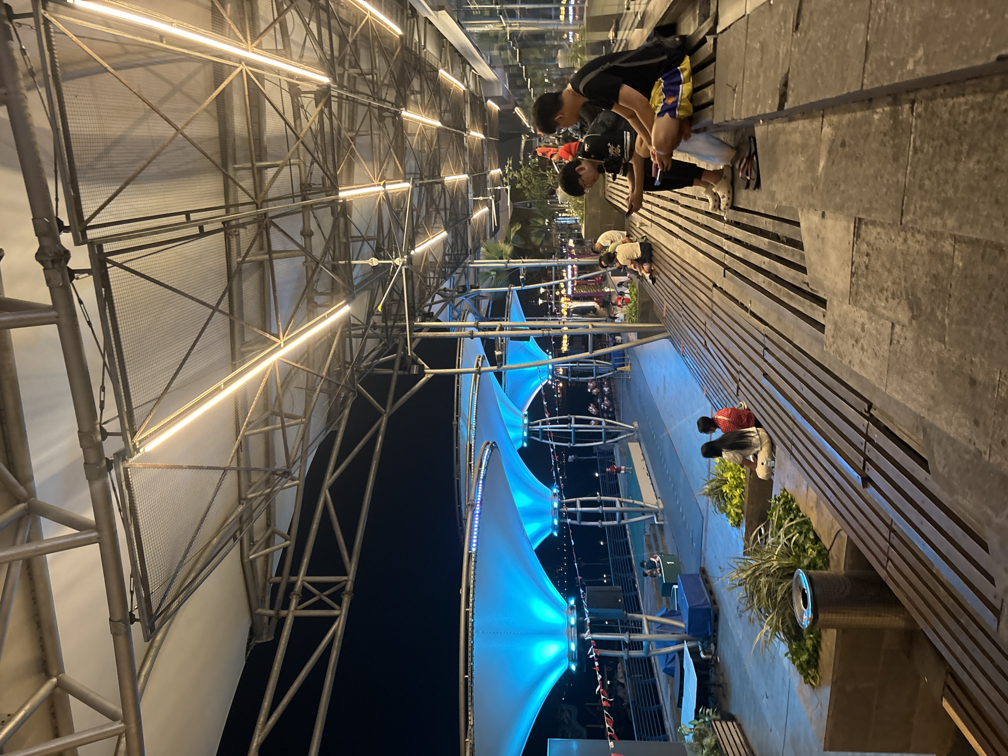 The image depicts an outdoor area at night, likely a public space like a waterfront promenade or plaza, possibly located in North Jakarta, Indonesia, given the context provided. The scene is illuminated by artificial lighting, with rows of bright, yellow lights running along the ceiling structure and prominent blue lighting under the translucent canopies in the background.
In the foreground, two young men are seated on a slatted wooden bench. The man on the left, wearing glasses and a black shirt with a white design, is looking at his phone. The man on the right is wearing a black t-shirt and shorts with yellow and blue accents, and he is also looking at his phone. Both are wearing casual footwear.
Further back, on a similar wooden seating structure, several other individuals are present. A group of three people, two women and one man, are sitting on a raised planter box with lush greenery. The two women are facing away from the camera, and one is wearing a red and white patterned shirt. Behind them, other figures are visible in the distance, seated or walking.
The architecture features a prominent metal framework supporting a stretched fabric roof, creating sheltered areas. In the background, large, illuminated blue structures, possibly awnings or shade sails, are supported by vertical poles. Beyond these, a body of water is visible, with lights reflecting on its surface, suggesting a harbor or river. There are also distant city lights and a dark night sky. A trash receptacle is situated near the planter box in the mid-ground. The overall environment suggests a recreational or dining area designed for public use.