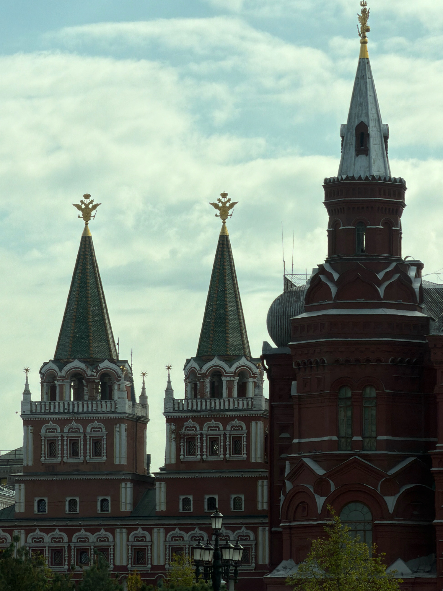 The image depicts a section of the Moscow Kremlin, specifically featuring several of its iconic towers and adjoining structures. In the foreground, three prominent towers with pointed, green-tiled roofs topped with golden double-headed eagles are visible. The tower on the far right is taller and appears to be part of a larger red brick building, likely the State Historical Museum, which is also visible with its intricate red brickwork and decorative elements. The sky is overcast with a pale, cloudy appearance. The buildings display a distinct architectural style characterized by red brick facades, arched windows, and ornate detailing. The overall scene captures a segment of Moscow's historical and architectural landscape under a subdued sky.