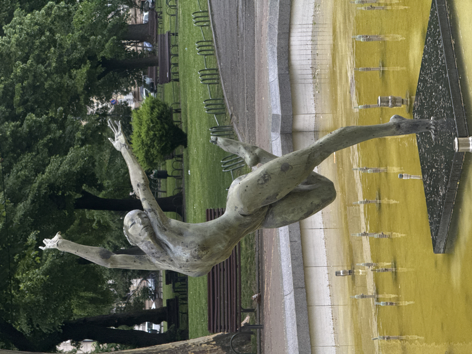 The image shows a bronze sculpture of a human figure in a pose that appears to be suspended or floating, seemingly emerging from a fountain. The sculpture is located in a park in the city of Sofia, Bulgaria.
The sculpture is positioned in the fountain, which has a geometric design and water of a yellow tint. The sculpture is the central element, and it stands in stark contrast to the natural green background of trees and grass, as well as some benches and bicycle racks in the park. The scene is captured on a seemingly overcast day.
