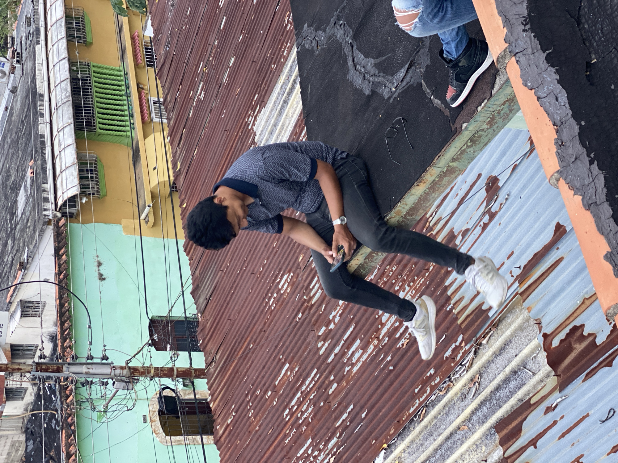 A medium, eye-level shot shows a man sitting on a corrugated metal roof, with parts of two buildings and two more people visible in the shot, taken in Tinaquillo, Venezuela.
The man in the foreground is Black, with dark curly hair. He wears a blue-patterned shirt with a dark blue collar and trim, dark jeans, and white sneakers. A silver watch is visible on his left wrist as he uses a cell phone.
The roof is a patchwork of corrugated metal in various colors and conditions. In the upper right of the frame, there is another person only partially visible, wearing light blue jeans with a hole in the right knee and black high-top sneakers with white soles. Another person's legs and black high-top sneakers are visible in the top right of the shot.
In the background, there are two buildings. One building has a light green exterior, and the other is yellow. The shot also shows several overhead power lines and the edge of a white building in the left corner.