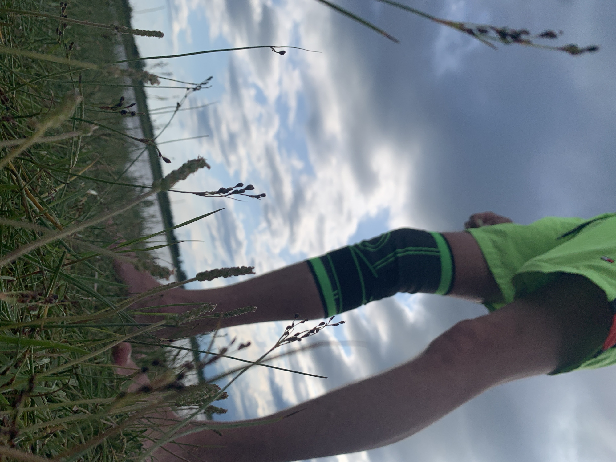 The image is a low-angle shot of a person's legs from the knee down, standing in tall grass near a body of water. The person is wearing bright green shorts and a black and green knee brace. The sky is cloudy with patches of blue. The foreground is filled with blades of grass and small plants, some with seed heads. The water is visible in the background, with a faint shoreline. The scene appears to be outdoors in a natural setting, possibly a park or embankment. The location is identified as Severodvinsk, Russia, implying this is a view of the coastline or a riverbank in that city.