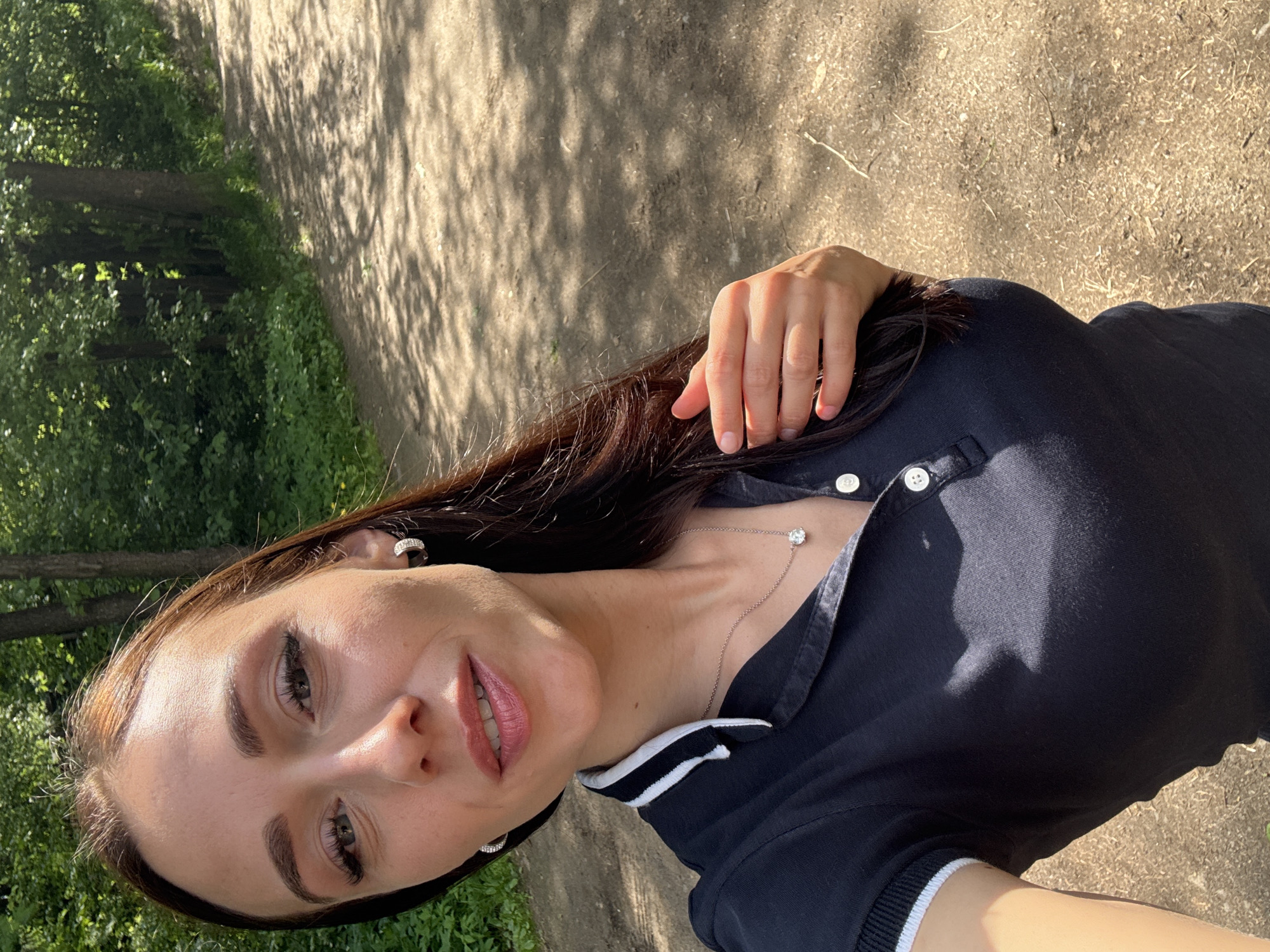 A young woman takes a vertical selfie outdoors in Sertolovo, Russia. She has long, dark brown hair, light skin, and a gentle smile, with visible makeup including defined eyebrows, mascara, and pink lip gloss. She wears a dark navy blue polo shirt with white buttons and distinctive white and black striped trim on the collar and sleeve cuffs. Accessories include small, possibly rhinestone, hoop earrings and a delicate silver chain necklace with a single clear gem pendant. Her right hand is visible resting on her hair near her shoulder.
The background reveals a natural setting with a dirt or gravel path to her right, and lush green trees and dense foliage to her left and behind her, indicating a park or wooded area. Strong, directional sunlight casts distinct shadows of tree leaves on the path and partially across her shirt, suggesting it is midday or early afternoon on a clear, sunny day. The woman appears relaxed and pleasant. No visible text is present in the image.