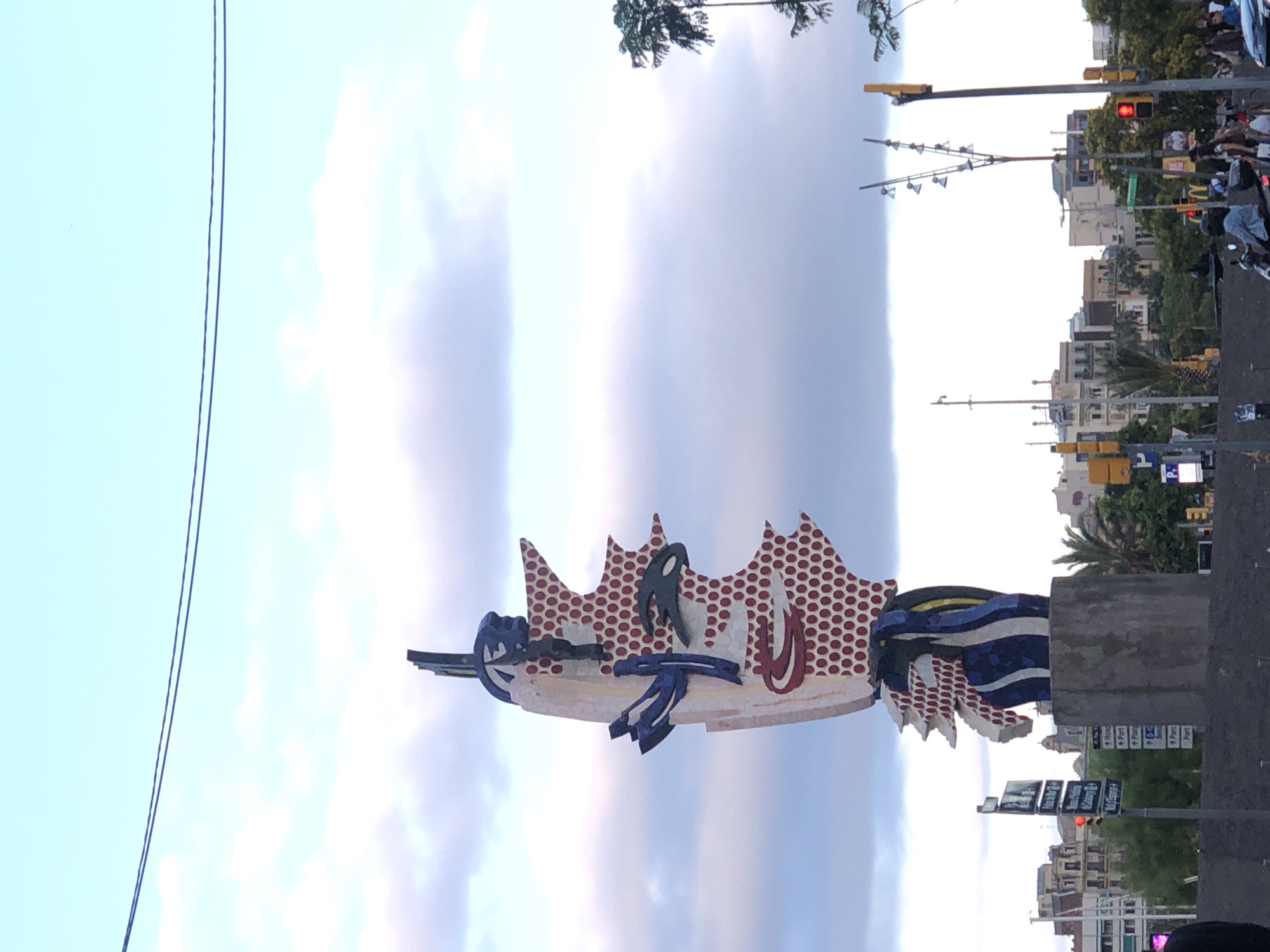 A large, colorful sculpture of a face stands in an urban setting, likely a city square or public park. The sculpture is abstract and features bold colors like red, blue, and white, with a pattern of red dots resembling a comic book style. It is placed on a grey concrete pedestal. In the background, there are buildings of various sizes and architectural styles, suggesting a city environment. Trees and other vegetation are visible, adding to the urban landscape. Traffic lights and street signs are also present, indicating a street or intersection. The sky is partly cloudy with a pale blue hue. Several people are visible in the lower part of the image, some walking, others on a scooter, suggesting a public space with pedestrian and vehicular traffic. The overall impression is a vibrant public artwork in a bustling city. The location is identified as Unknown, Unknown.