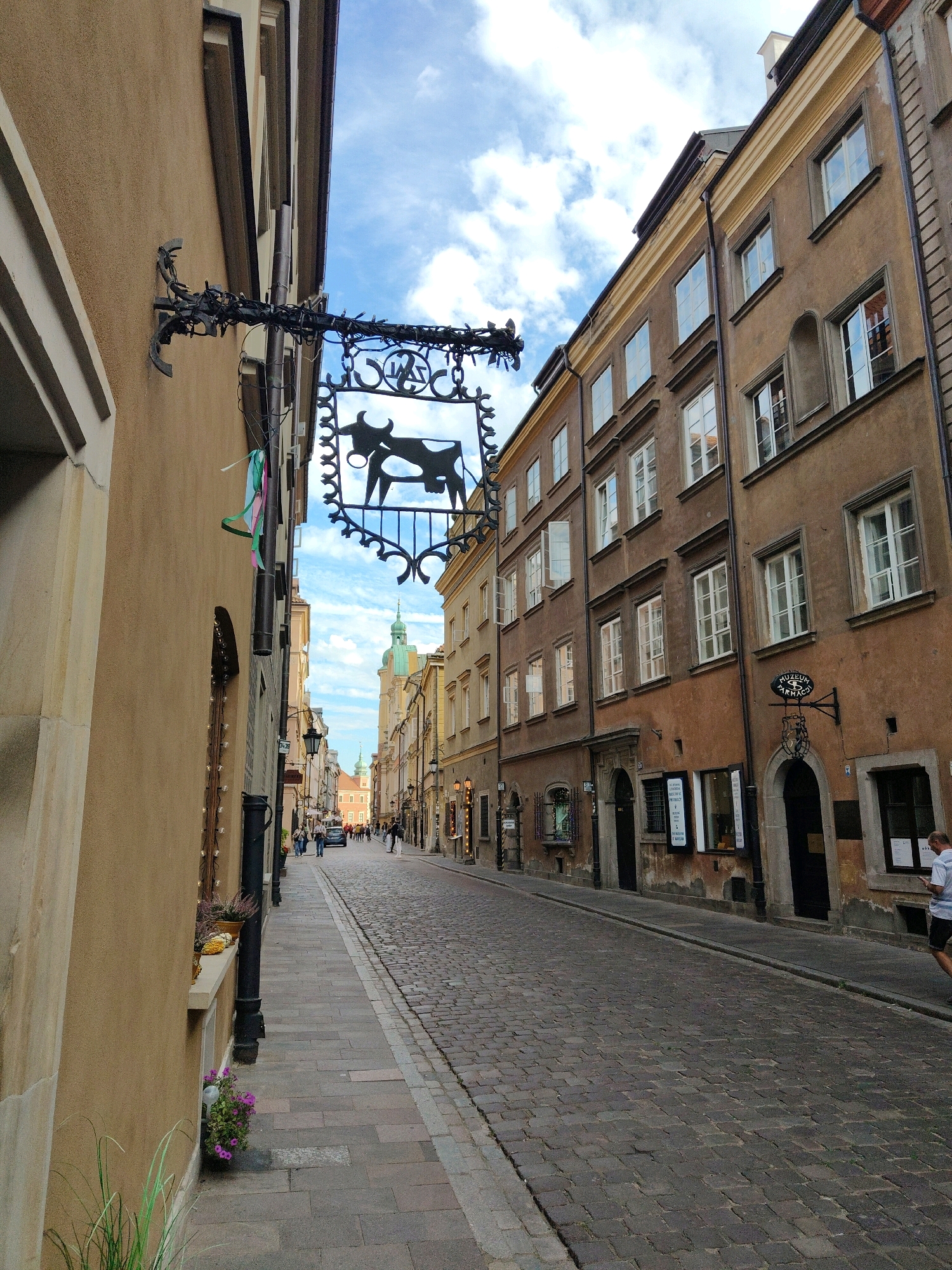 The image depicts a street scene in Warsaw, Poland. The perspective is looking down a cobblestone street lined with historic buildings on both sides. A prominent black wrought-iron sign with a cow silhouette hangs from an ornate bracket on the left side of the street, just below the eaves of a building. The sign is decorated with what appear to be small lights wrapped around the bracket. Colorful ribbons are also attached to the bracket.
The buildings are several stories high with a warm, muted color palette, featuring numerous windows, some of which are open. The architecture suggests an old European city. The street itself is paved with cobblestones. In the distance, a green-domed tower is visible, indicating the presence of a church or other significant landmark.
There are several people walking on the sidewalks and crossing the street. On the right side of the street, a man in a white shirt and dark shorts is walking away from the camera, looking at his phone. Other individuals are seen further down the street, appearing to be tourists or residents. The sky above is blue with scattered white clouds, suggesting it is daytime. The overall atmosphere is one of a well-preserved historic district in an urban environment.