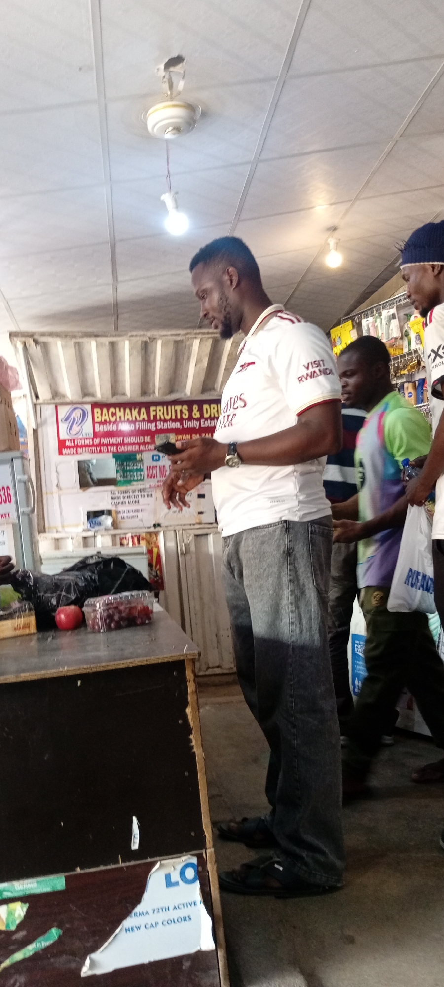 The image depicts an indoor scene, likely a market stall, with several people present. The primary subject is a man in the foreground, wearing a white and red-striped polo shirt and dark jeans. He is holding a smartphone in his hands and appears to be looking at it. To his right, other individuals are visible, including one wearing a dark beanie and another in a brightly colored shirt.
Behind the man, a stall is set up with various items, including what appear to be fruits and drinks, as indicated by the text on the signage. The signage reads "BACHAKA FRUITS & DRIVES" and provides instructions for payment. Other signs and labels are also visible, some of which are partially obscured or difficult to read. Objects on the counter include a plastic container of grapes, a red apple, and a black plastic bag.
The setting appears to be a somewhat utilitarian indoor space with a tiled ceiling and fluorescent lighting. The overall atmosphere seems casual and transactional. The time of day is not explicitly indicated, but the lighting suggests it's daytime. The location context provided is Aviation Quarters, Nigeria.