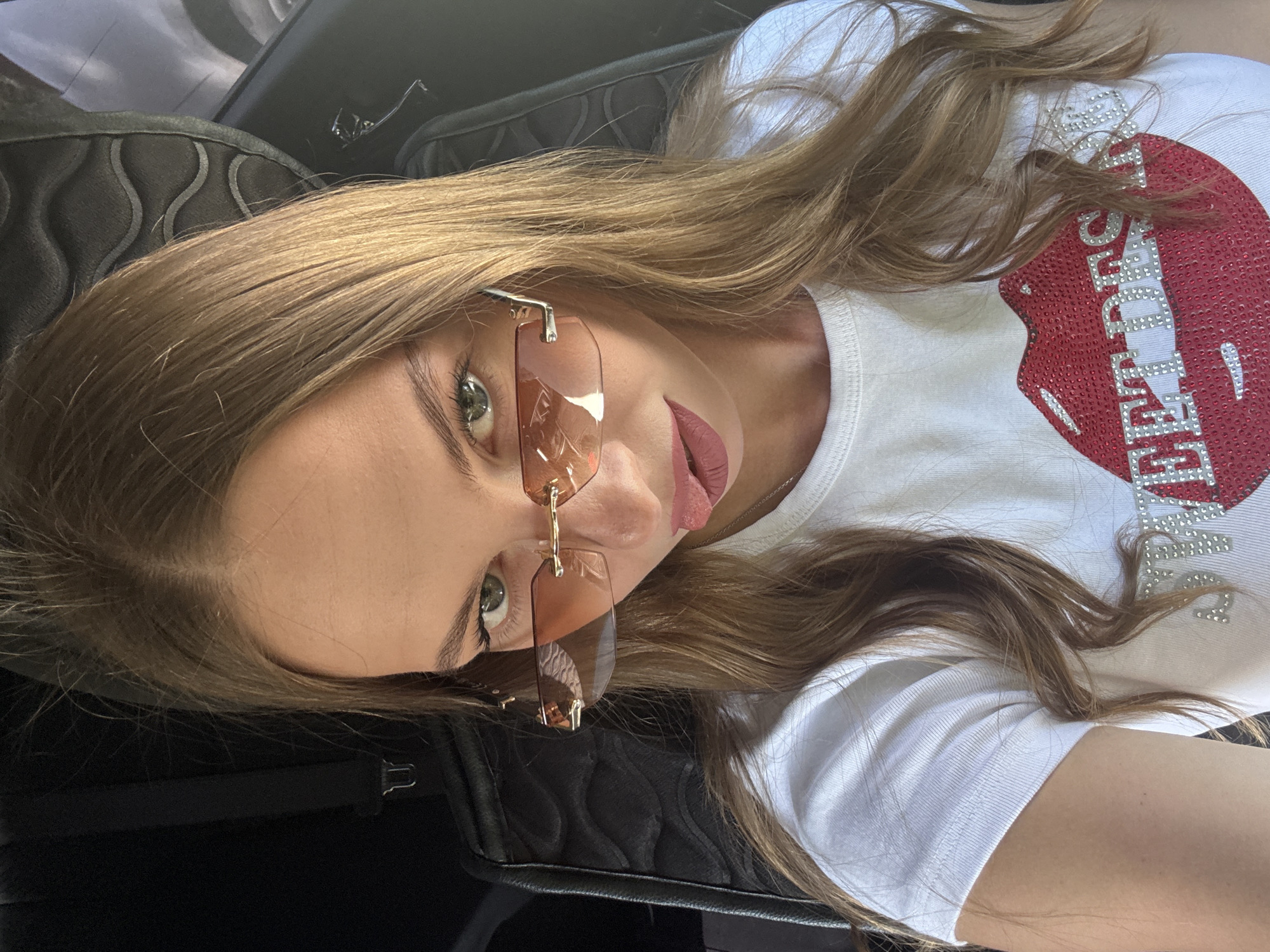 A young woman is captured in a close-up, vertical, low-angle shot, likely taken indoors. She has long, blonde hair that falls around her face and shoulders. She is wearing a white t-shirt with a large, sparkling red heart and the word "SWEETNESS" emblazoned across it in silver rhinestones. Her eyes are visible through rimless, square, tinted sunglasses. She is also wearing lipstick. Her skin appears smooth and fair. The background consists of dark, textured upholstery, possibly from a car seat or a chair. The overall mood appears casual and perhaps a bit playful, with the woman's gaze directed upwards and slightly away from the camera. The lighting suggests it is daytime.