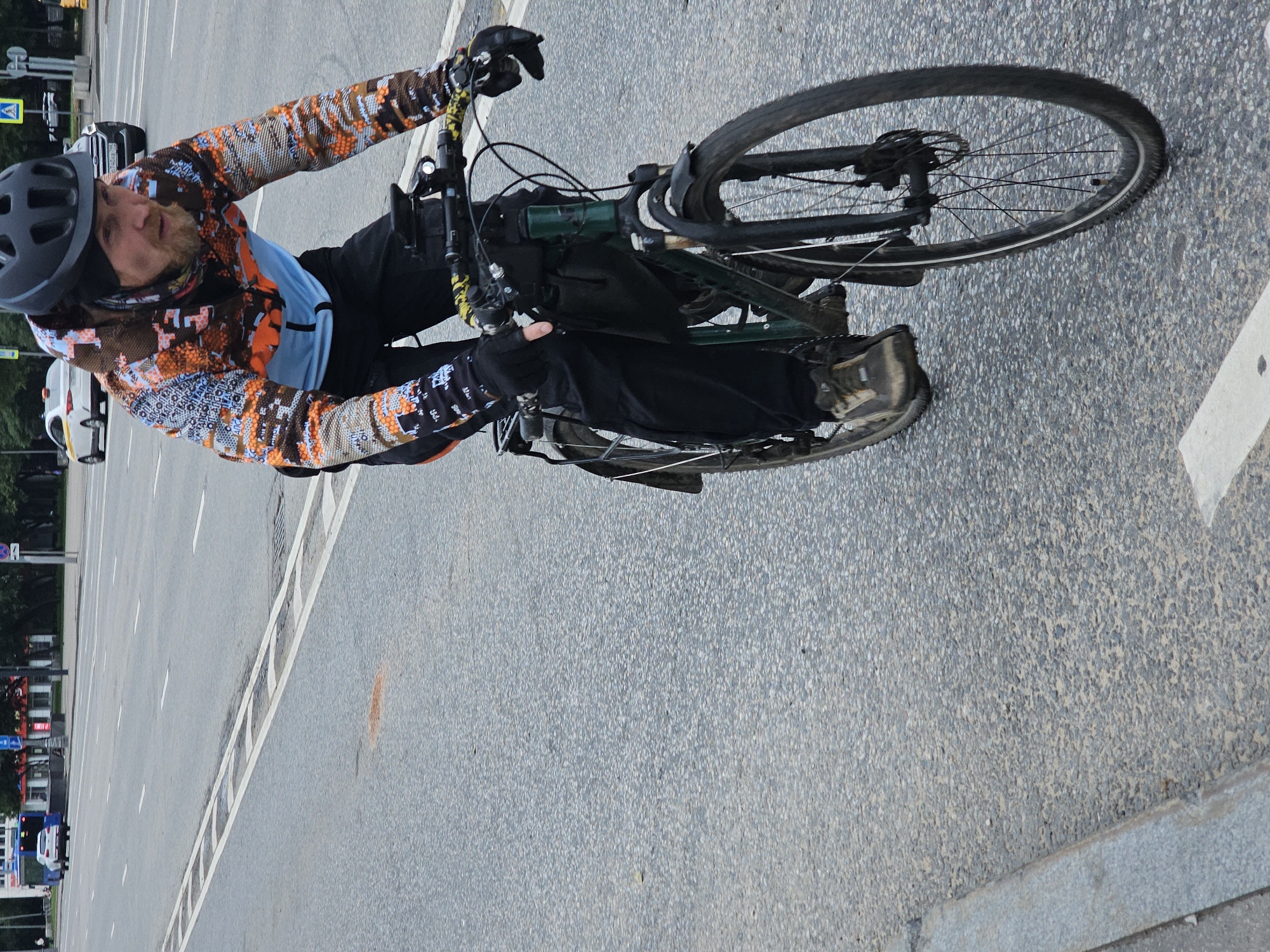 A man wearing a black helmet and a patterned jacket with orange, blue, and brown colors rides a dark green bicycle on an asphalt road in Moscow, Russia. He is also wearing black pants and black gloves. The road has white lane markings. In the background, there is a white car, a yellow taxi, and buildings. The man is in motion, pedaling the bicycle.