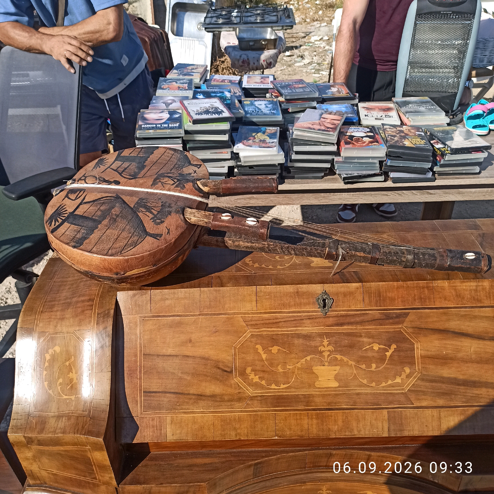 This image depicts an outdoor market scene in Sannicandro di Bari, Italy, on September 6, 2026, at 9:33 AM. A person, partially visible and wearing a blue short-sleeved shirt and dark shorts, is leaning on a black office chair. In the foreground, an antique wooden furniture piece with intricate inlay work is prominently displayed. On top of the furniture, a stringed musical instrument with a circular wooden body decorated with black tribal-like patterns is placed. Several stacks of DVDs are arranged on a shelf or table behind the musical instrument. In the background, a portable gas stove and a heater are visible. A pair of turquoise and pink flip-flops are also present on the right side of the frame. The overall environment suggests a sunny day at a street market or flea market.