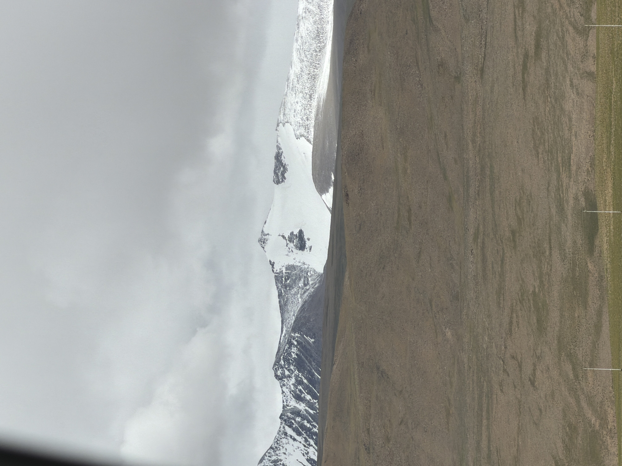 The image depicts a vast, arid landscape with snow-capped mountains in the background. The foreground is a barren, brownish-green plain, sparsely dotted with vegetation and a few white vertical posts that appear to be part of a fence or power line. The mountains, partially covered in snow, rise majestically against a cloudy, overcast sky. The overall impression is one of a remote, high-altitude environment. The scene is devoid of human presence.