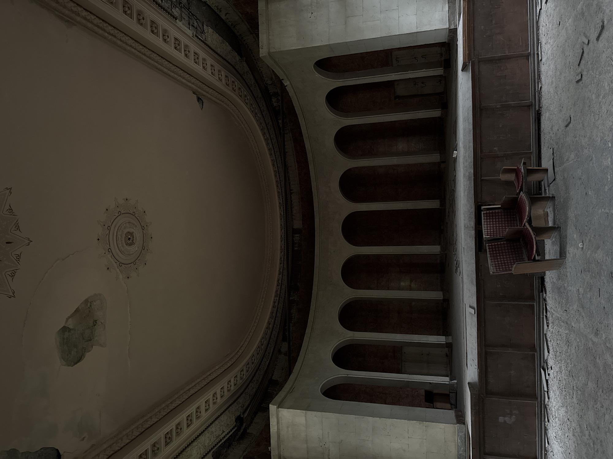 The image depicts the interior of an abandoned theater in Tskaltubo, Georgia. The frame is oriented from a low angle, looking up at the ceiling, mezzanine level, and side wall.
The ceiling is a large, oval dome with ornate decorative moldings and a central medallion. It shows signs of significant water damage and deterioration.
Adjacent to the ceiling is a mezzanine level with a series of arched openings supported by columns or pillars. Behind the arches, glimpses of the dark interior can be seen.
On the right side of the image, a portion of the side wall is visible, constructed from gray stone or concrete. Attached to the wall are several rows of theater seating upholstered in a faded red and black checkered pattern. The wall shows signs of weathering and neglect.
The overall environment appears decayed and neglected, consistent with an abandoned or disused space. No people are present in the image.