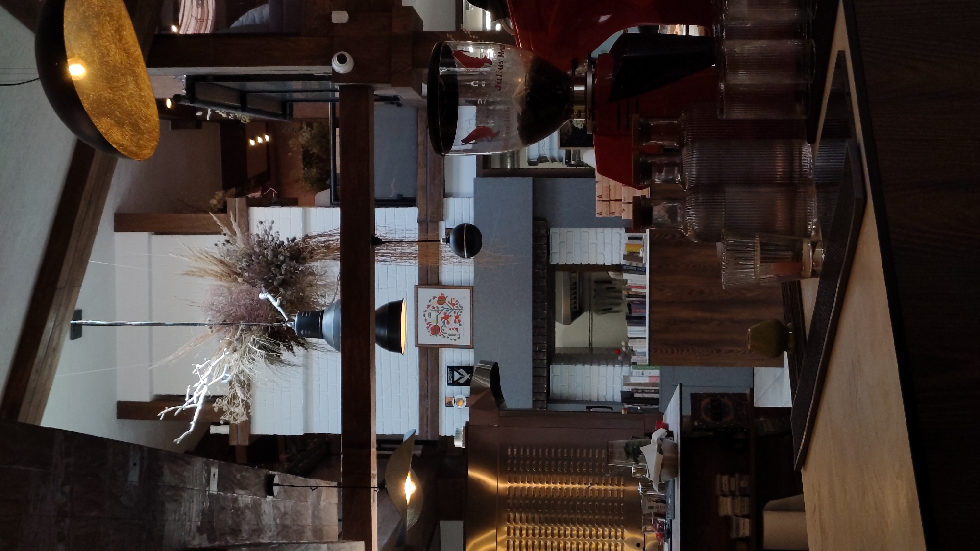 The image depicts the interior of a cafe, possibly located in Plovdiv, Bulgaria, characterized by a rustic and modern aesthetic. Dominating the foreground is a polished wooden counter, with a black matting surface and a reflective dark rectangular object placed on it. Behind this, several glass bottles filled with clear liquid stand alongside fluted glasses, and a coffee grinder with a clear hopper filled with coffee beans is visible.
Further back, a metal appliance, possibly a commercial oven or refrigerator, is positioned to the left. A wall with exposed brickwork is visible behind it, with shelves displaying books and decorative items. A framed artwork featuring floral motifs hangs on the wall.
Overhead, industrial-style pendant lights, some with exposed bulbs and others with dark shades, illuminate the space. Decorative dried floral arrangements hang from the ceiling. The overall atmosphere suggests a contemporary cafe with elements of natural materials and industrial design. No individuals are directly visible in the frame.