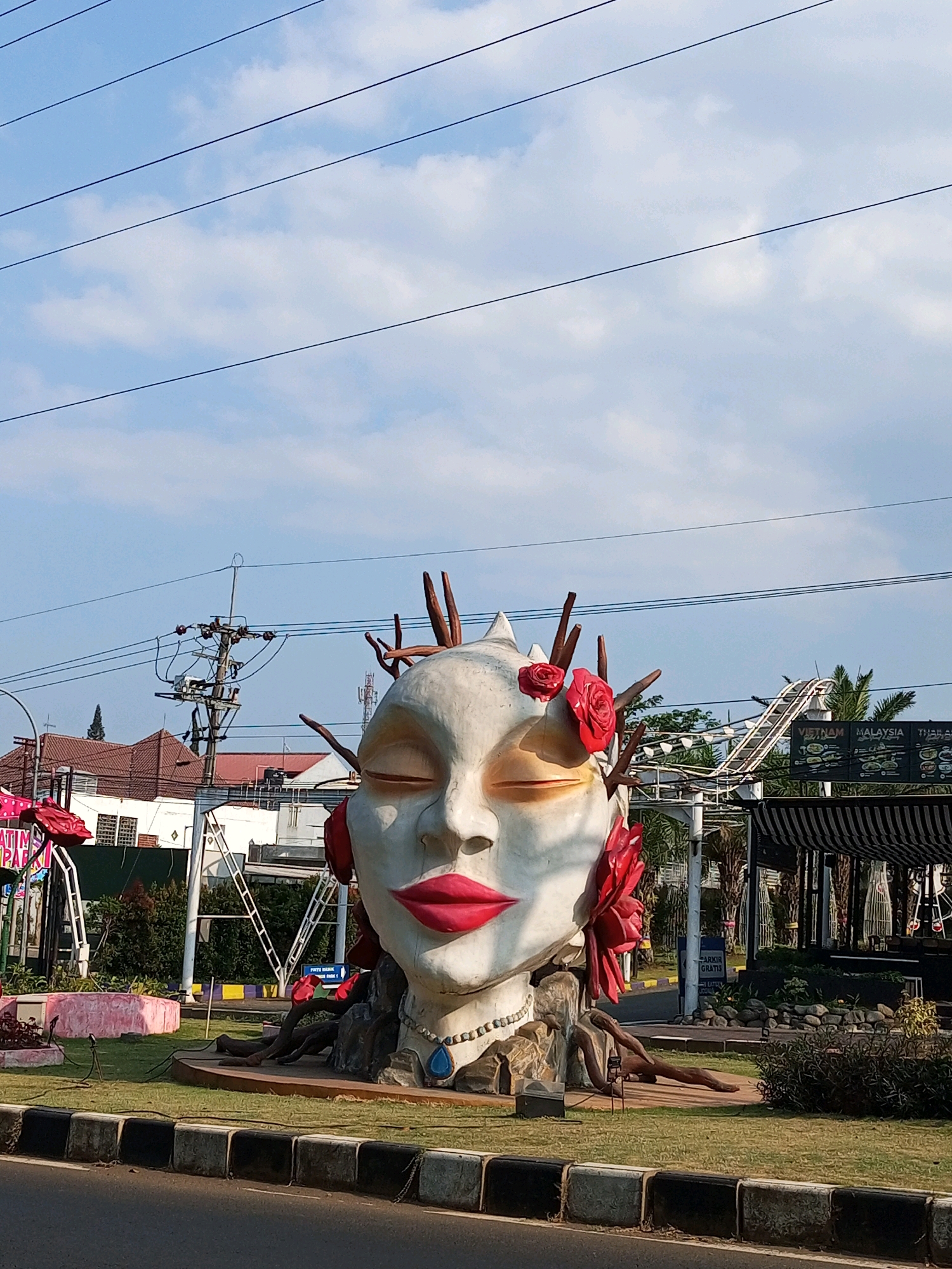 A large, white sculpture of a stylized female head with closed eyes, bright red lips, and red roses adorning its hair stands in a grassy roundabout in front of a street in Batu, Indonesia. The sculpture has a serene expression, and its base is adorned with rocky elements and exposed roots. To the right of the sculpture, a roller coaster track is visible against a blue sky with scattered clouds and overhead power lines. A striped awning and a sign indicating "PARKIR GRATIS" (Free Parking) are also present. The foreground shows a paved road with a concrete divider. The background features buildings with red-tiled roofs, trees, and utility poles.