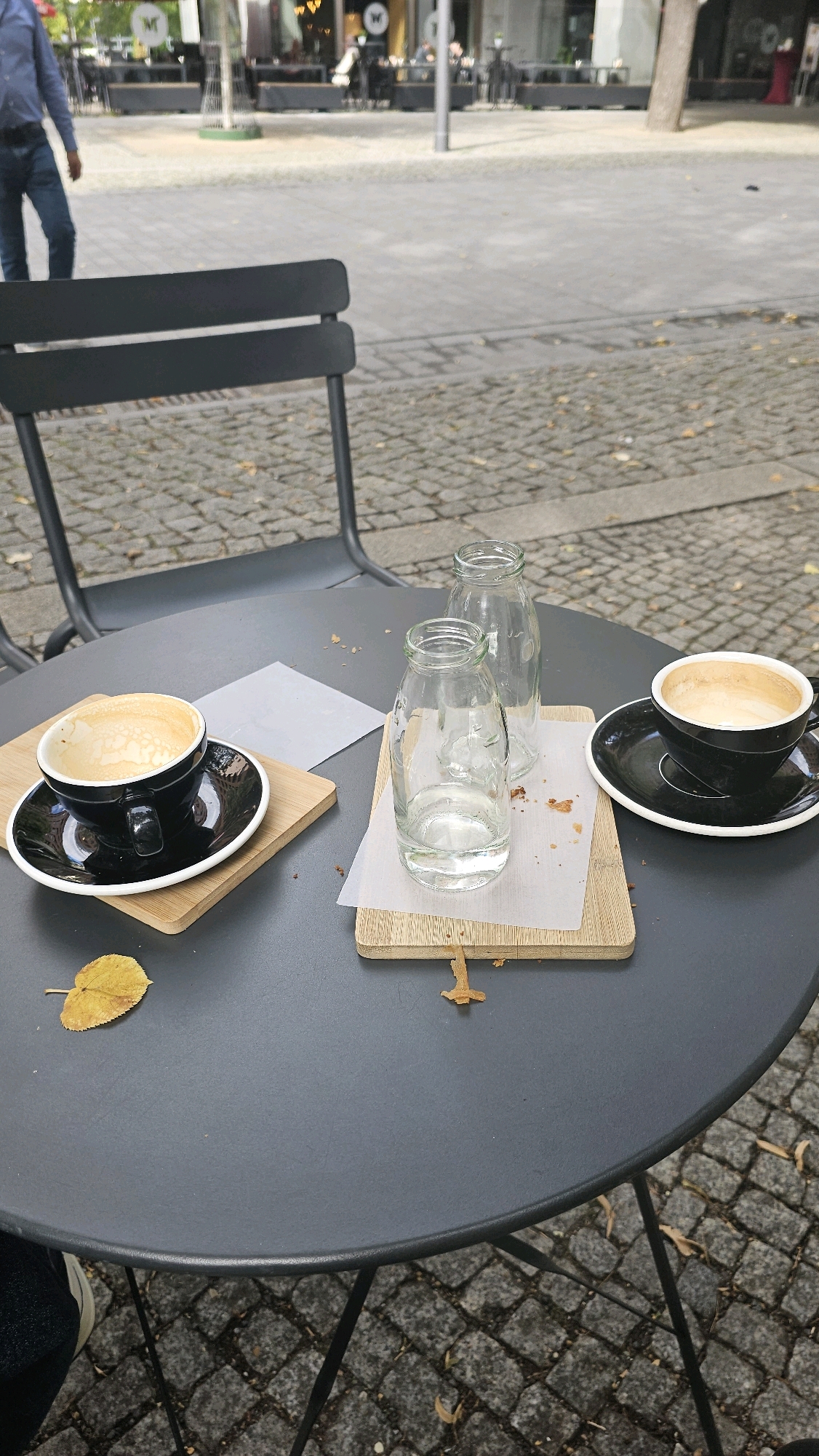 An outdoor cafe scene is depicted in Berlin, Germany, on a cobblestone walkway. A dark gray, round metal table occupies the foreground, with two chairs visible behind it. On the table, two black coffee cups sit on saucers. The cup on the left contains a frothy latte, while the cup on the right has a nearly empty latte. Two clear glass bottles, one full of water and the other empty, are placed on a wooden board. A single yellow leaf rests on the table near the left cup, and other fallen leaves are scattered on the ground. In the background, a person in a blue shirt and jeans walks away from the camera. A blurred urban environment with buildings and trees can be seen further in the background.
