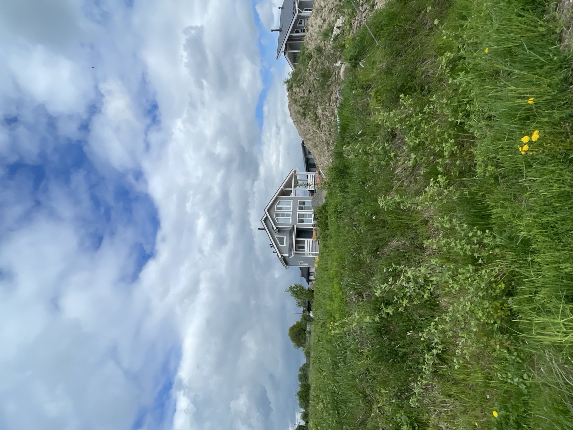 The image shows a two-story gray house with white trim, situated in Solov'evka, Russia. The house is angled against a background of a cloudy sky with patches of blue. To the right of the house is a grassy slope with wildflowers, and another house is partially visible on top of the slope. The grass appears green and untamed. The overall scene suggests a rural or suburban setting.