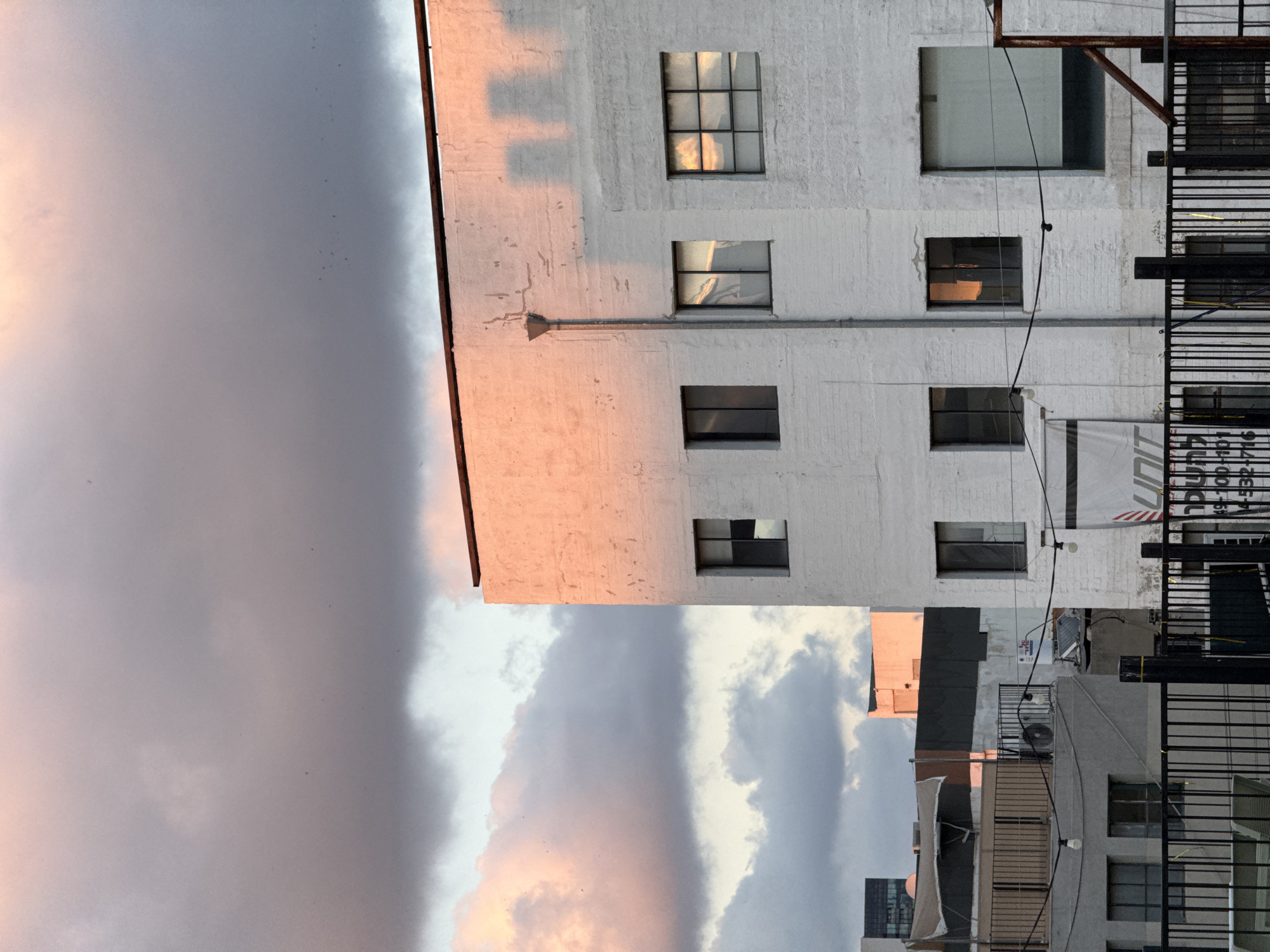 The image shows an urban scene, presumably in Tel-Aviv, Israel, given the architectural style and general environment. The sky takes up a large portion of the frame, displaying a mix of dark gray storm clouds and patches of lighter, sunset-lit clouds. A tall, white-painted building with multiple windows dominates the right side of the image. The building appears to be multi-storied, with a fire escape attached to its side. The windows on the building reflect the warm colors of the setting sun. To the right of the white building, a series of other structures can be seen, including a gray-colored building with balconies and air conditioning units. Power lines crisscross the area. A banner or sign is partially visible, attached to the white building. The image is taken from a low angle, looking upwards.