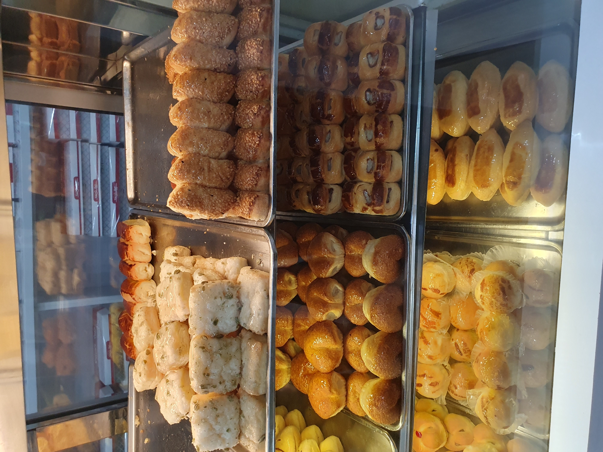 The image shows a display of baked goods in a bakery, likely in Shiraz, Iran, given the context. The display is organized on multiple shelves within a glass-fronted cabinet.
The top shelf, visible on the left, contains white, rectangular pastries topped with a glossy glaze and what appear to be small, green pieces, possibly herbs or seeds. Behind these, and slightly out of focus, are rows of similar pastries and some golden-brown baked goods.
The next shelf down, also on the left, features a tray filled with golden-brown, rounded pastries that are slightly flattened on top. Some of these have a visible split or indentation. To the right of this shelf, and extending to the full width of the display, is another shelf holding rows of elongated pastries. These pastries are covered in sesame seeds and have a golden-brown crust.
The shelf below that contains rows of small, round pastries with a dark filling visible through a slit on their surface. These are arranged neatly in trays.
The bottom shelf, on the right side of the display, shows a tray of oval-shaped pastries with a lighter, golden-brown top that has a slightly darker, rectangular or square marking. These pastries are also glazed.
The overall environment appears to be a commercial setting, a bakery or pastry shop, with the baked goods presented for sale. The lighting suggests an interior setting, likely under artificial lights. The cleanliness of the display cases and trays indicates a maintained commercial space.