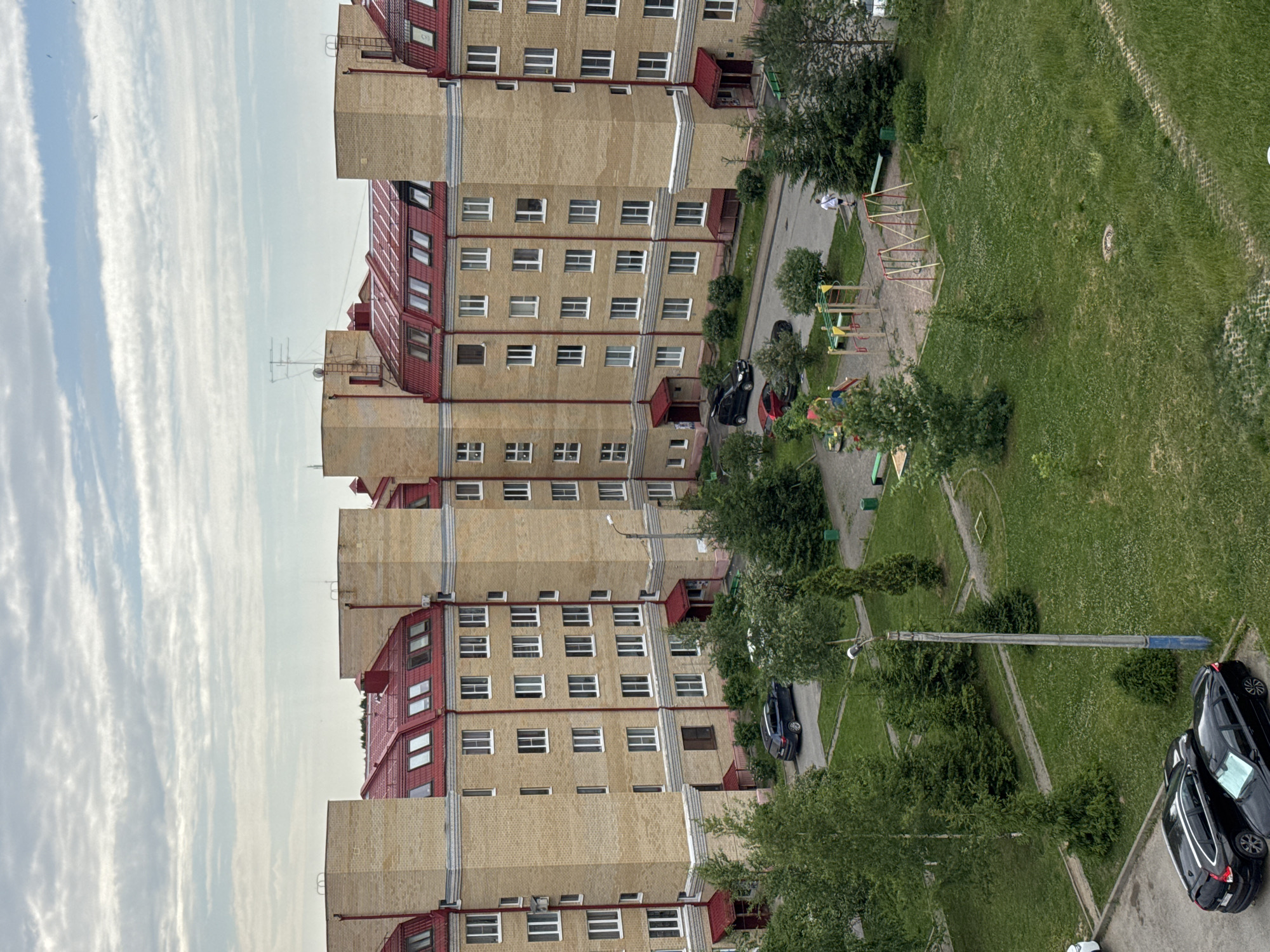 An aerial view captures a modern residential apartment complex in the city of Kirishi, Russia. The building, constructed with light yellow bricks and featuring distinctive red, mansard-style roofs with dormer windows, forms a U-shape around a central courtyard. The courtyard is a vibrant green space with mature trees and a children's playground equipped with swings and climbing structures. Several vehicles are parked on asphalt surfaces surrounding the complex and within the courtyard. The sky above is filled with a dramatic pattern of grey and white clouds, suggesting a cloudy or overcast day. A single person is visible walking in the courtyard.