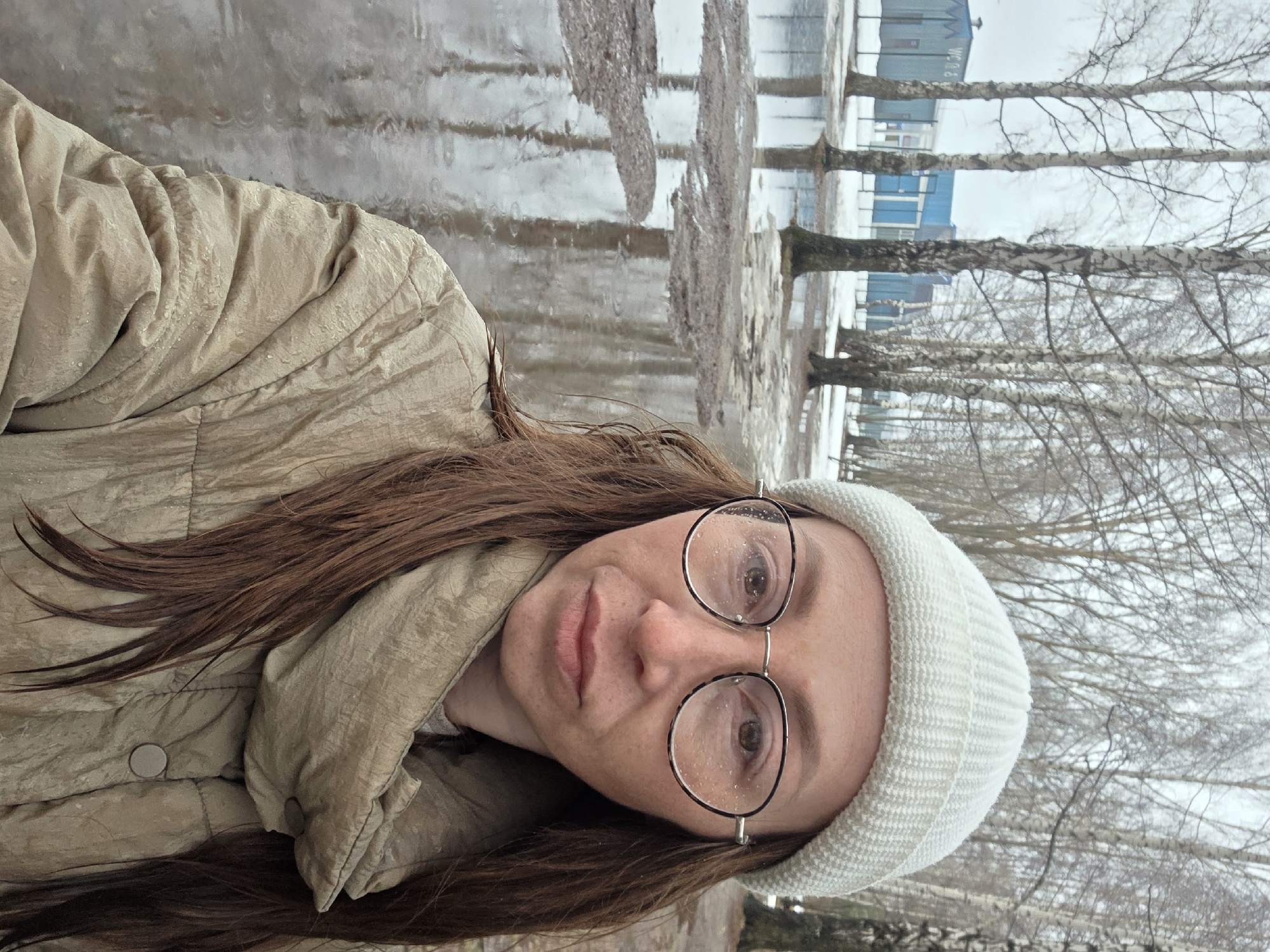A woman wearing a beige jacket and a white beanie is looking at the camera. She is wearing glasses with water droplets on the lenses. The background features a landscape with puddles and birch trees. In the distance, a building with blue accents is visible. The location is Chaikovsky, Russia.