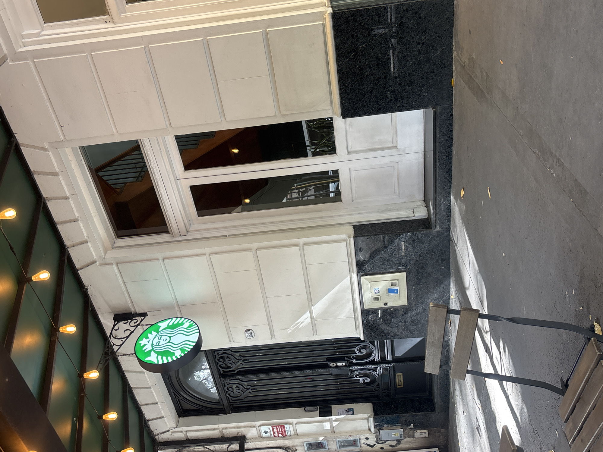 A Starbucks location in Buenos Aires, Argentina, is viewed from below. The building facade is primarily white, accented with black marble. The Starbucks logo, green and round, is visible. String lights hang along the side of the building. Two windows are visible, reflecting interior features. An ornate black gate covers the entrance. A chair sits on the sidewalk in front of the building.