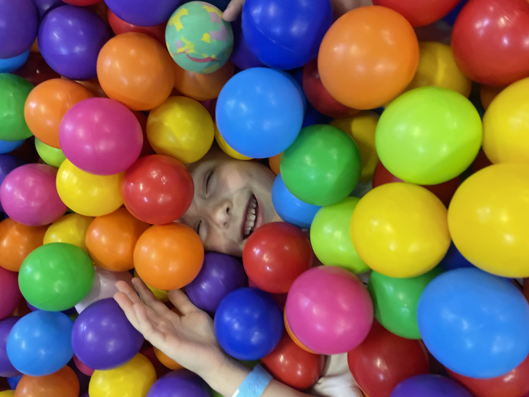A child lies amidst a dense collection of multicolored plastic balls, primarily red, orange, yellow, green, blue, and purple. The child's face, with closed eyes and a smile revealing teeth, is partially visible among the balls. A hand is visible amidst the balls with a blue wristband. The setting appears to be an indoor play area. There is no definitive information to place it within Sochi, Russia.