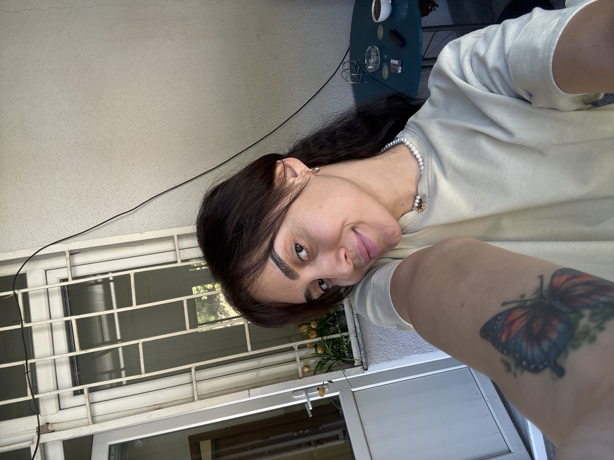 In Tbilisi, Georgia, a woman with dark hair and a butterfly tattoo on her arm takes a selfie outdoors. She is wearing a pearl necklace and a light-colored t-shirt. In the background, there is a window with bars, some yellow flowers in a planter, a white door, and a teal-colored table with a coffee cup, an ashtray, and a pack of cigarettes. The overall environment appears to be a patio or balcony.