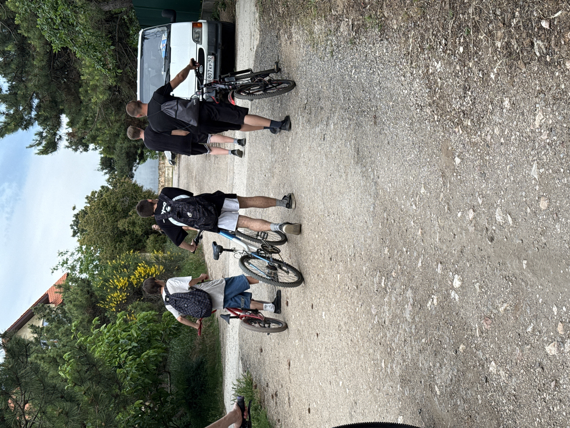 This image, taken in the city of Unknown, Unknown, depicts a group of five individuals walking along an unpaved, gravel road on an overcast day.
In the foreground and mid-ground, four young adult males are walking away from the camera, pushing bicycles. From left to right:
1. A person, largely out of frame on the far left, shows an arm and leg wearing light-colored shorts and sandals. Part of a bicycle's handle and front wheel are visible.
2. A male in a white t-shirt, blue denim shorts, white socks, and dark shoes, wears a dark backpack with a checkered pattern. He is pushing a small red BMX-style bicycle.
3. A male in a black t-shirt, black athletic shorts with white vertical stripes on the sides, white socks, and dark shoes. He is not pushing a bicycle.
4. A male in a black t-shirt, black shorts, and dark shoes, wears a grey drawstring backpack. He is pushing a dark-colored mountain bike with blue accents on the frame and black wheels.
Further in the background, to the right, a fifth, fainter figure, possibly female, in a light-colored top, is walking along the road.
The road surface consists of compacted dirt and varying sizes of light grey and reddish gravel. Sparse dry vegetation is visible along the road's edges.
On the right side of the road in the mid-background, a white Ford Transit-style van is parked, facing away from the camera. Its license plate, "E 807 KP 92," is clearly visible.
The surrounding environment features dense green foliage, including tall pine-like trees and various shrubs. On the left side of the road, behind the first two individuals, yellow flowering shrubs are prominent. A two-story building with a red-tiled roof is partially visible through the trees on the left. The sky is largely overcast with light grey clouds.
