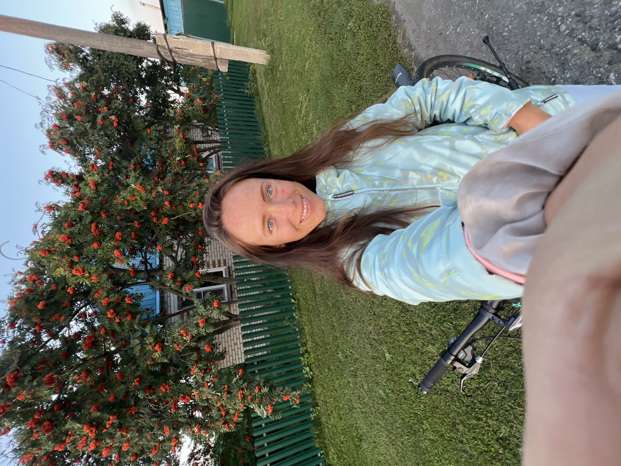 A woman with long brown hair and blue eyes is taking a selfie outdoors in a rural area. She is wearing a light blue, patterned jacket and is standing next to a bicycle. Behind her is a green fence, a house with a blue roof, and a tree laden with bright red berries. The setting appears to be a yard or garden in Pivkino, Russia. The sky is clear and blue, suggesting it is daytime. The woman is smiling and looking at the camera, holding her arm out to capture the photo. The overall scene conveys a relaxed and pleasant atmosphere.