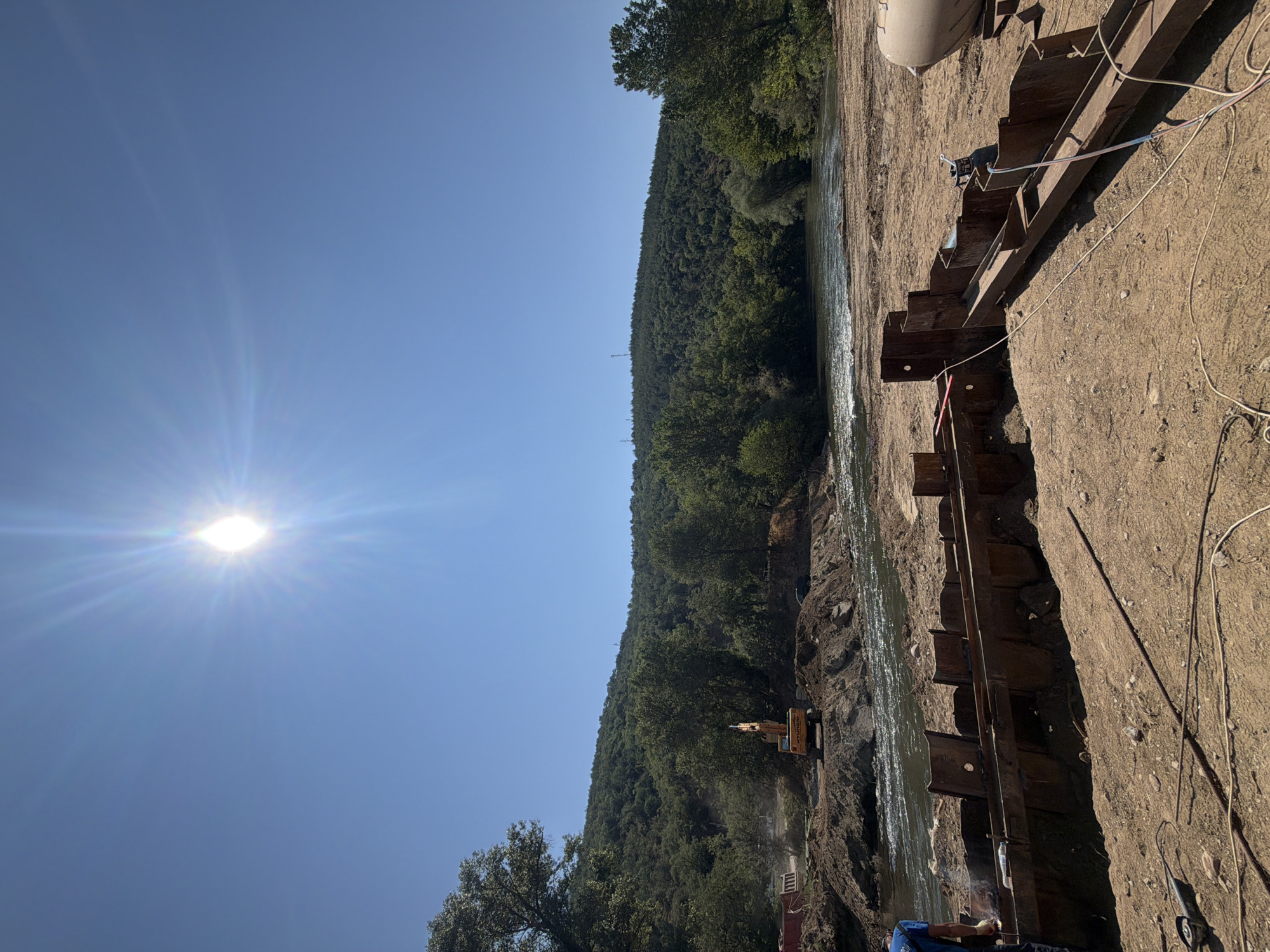 The image shows a construction site by a river, likely in Kresna, Bulgaria. In the foreground, metal sheet piling is being installed on a sandy embankment. A person is visible on the left, operating a welding torch, indicating active construction. Various construction tools and materials, including cables and a grinder, are scattered on the ground. In the middle ground, an excavator is seen working on the riverbank, with piles of earth and debris. The river itself is flowing, with sunlight reflecting off its surface. The background features a lush, tree-covered hillside under a clear blue sky with a bright sun. The overall scene depicts an active construction project in a natural riverside environment.
