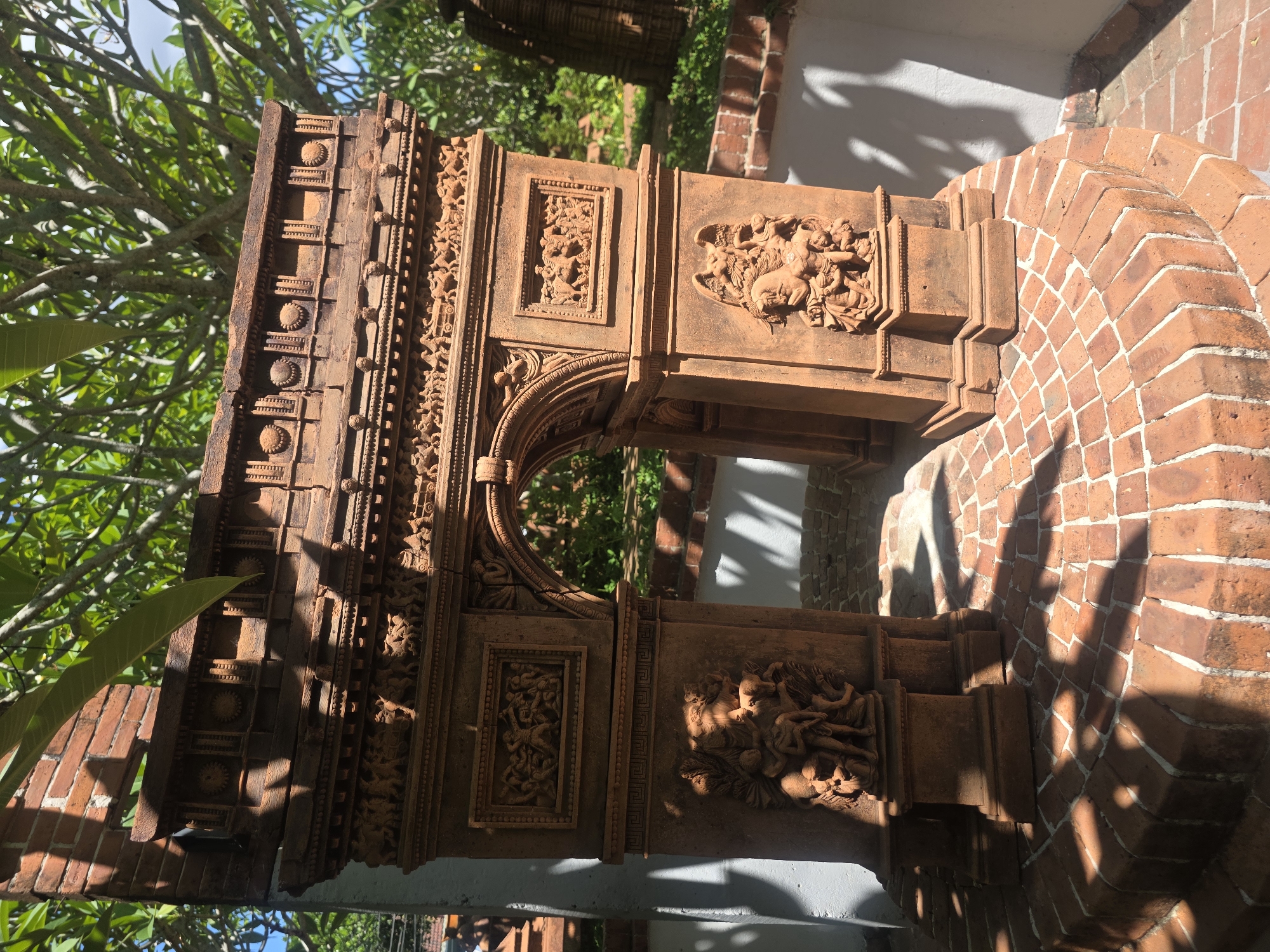 The image displays a section of an intricately carved architectural structure, likely made of terracotta or dark wood, set outdoors in Đà Nẵng, Vietnam. The reddish-brown structure features an ornate archway or gateway adorned with detailed relief carvings.
The carvings include large panels depicting multiple figures, possibly mythological beings or humans engaged in a scene, and smaller framed sections with equally elaborate, densely packed figures. The top and horizontal beams of the structure are decorated with repetitive patterns, including geometric shapes and circular motifs resembling rosettes.
To the right of the carved element, a brick archway with red-orange bricks laid in a decorative pattern (possibly herringbone) is visible, partially in shadow. Lush green foliage from trees and bushes frames the background and foreground, suggesting a natural or garden setting.
The scene is illuminated by bright sunlight, casting strong, well-defined shadows across the brickwork and the lower parts of the carved structure, indicating either morning or late afternoon. The clear weather and vibrant natural light highlight the textures and depth of the ancient craftsmanship. No visible text is present in the image.