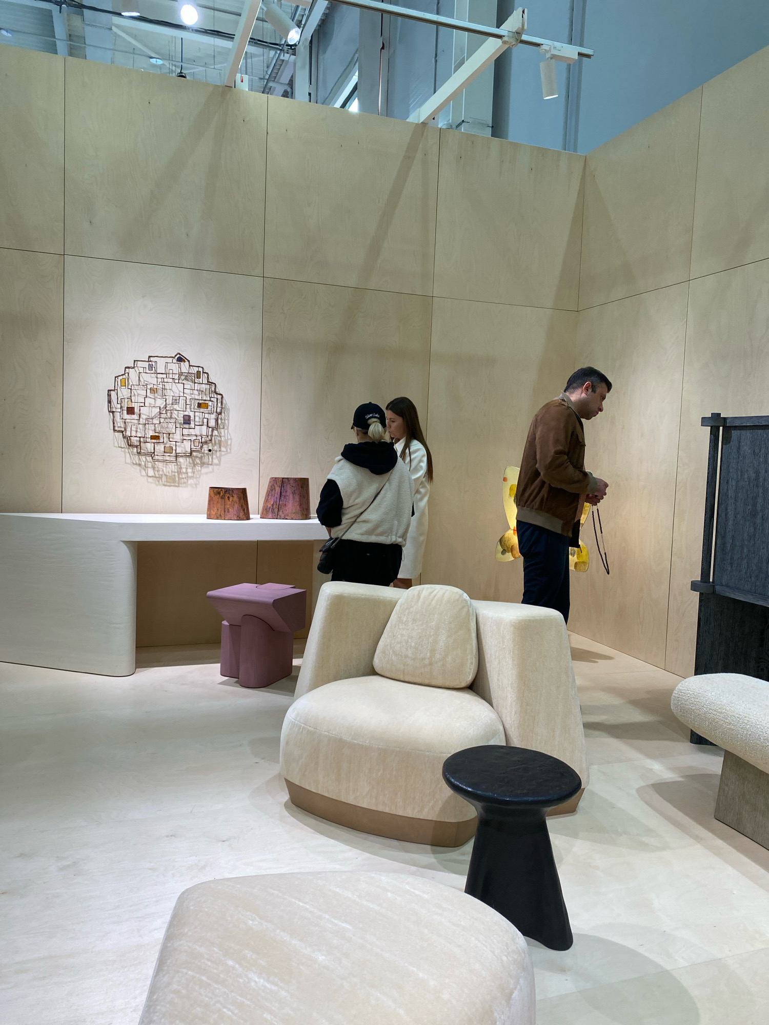 The image depicts an indoor exhibition space, possibly at Crocus Expo, a convention center located in Krasnogorsk, Moscow Oblast, Russia, near the MKAD beltway bordering Khimki. The environment is sparsely decorated with light-colored wood paneling on the walls and floor.
In the foreground, a large, plush, cream-colored armchair with a beige base is positioned slightly off-center. To its right and slightly behind, another cream-colored seating element, possibly an ottoman or another part of a modular sofa, is visible. In front of the armchair is a small, black, stool-like side table with a flared base and a rounded top.
To the left, a long, curved, white counter or desk with a matte finish dominates the middle ground. On top of this counter, there are two cylindrical decorative objects with a textured, reddish-brown surface and a smaller, irregularly shaped, pink stool with four legs. Above the counter, a circular wall art made of metal and glass inserts is mounted.
Two women are standing by the counter, facing the wall art and discussing it. One woman, wearing a black baseball cap and a beige and black jacket, is closer to the viewer. The other woman, with long brown hair, is wearing a white top.
Further to the right, a man in a brown jacket and dark pants is standing and looking at a decorative yellow object with a butterfly-like shape, which is mounted on the wall. He is holding a beaded necklace in his hands. Adjacent to him and on the right edge of the frame is a tall, dark-colored cabinet or display unit.
The lighting in the space appears to be overhead spotlights, creating a bright and airy atmosphere. The overall impression is of a contemporary design or art exhibition.