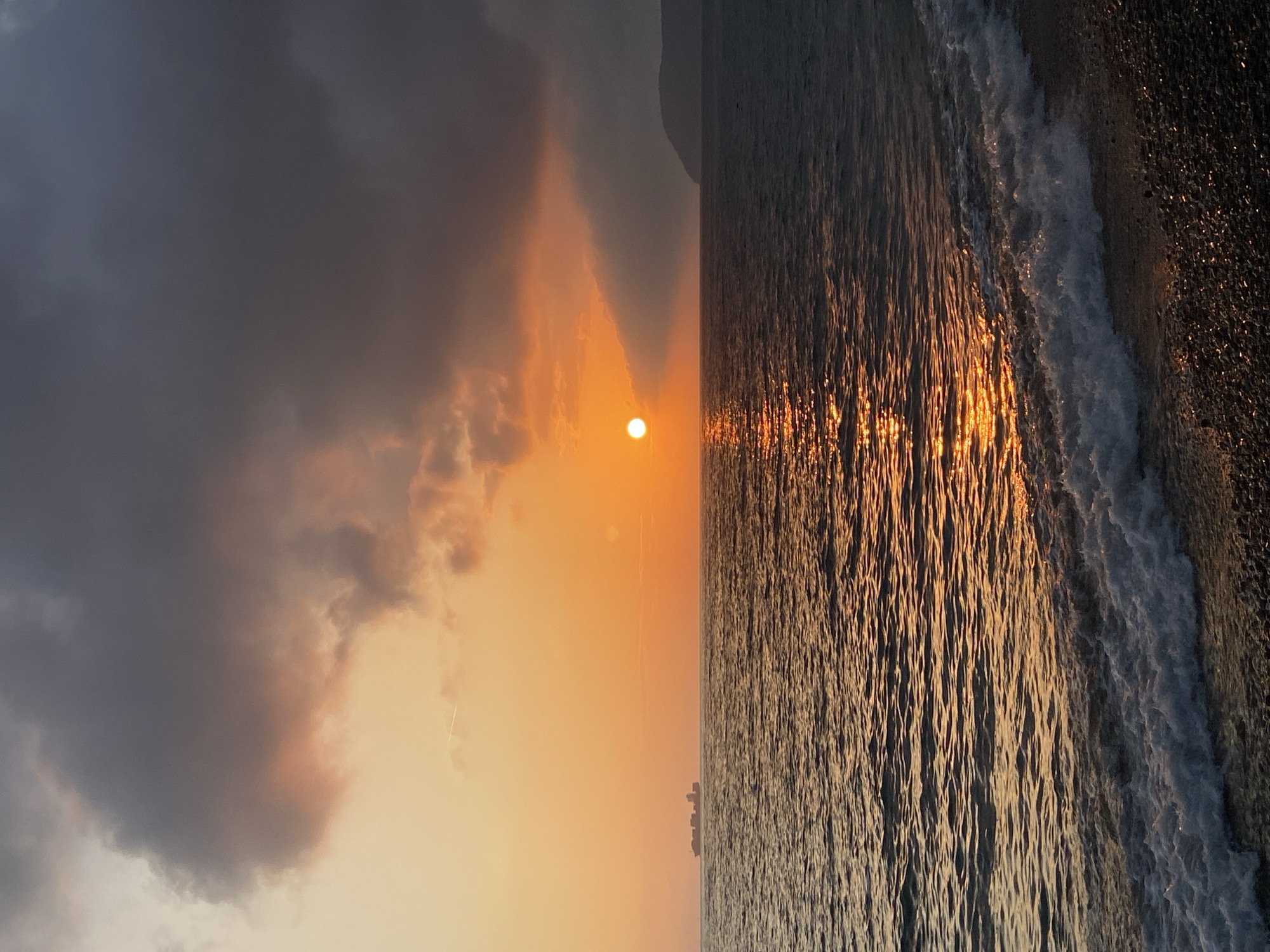 The image depicts a sunset over the Adriatic Sea in Bar, Montenegro. Dark, ominous clouds fill the left portion of the sky, while the sun peeks through a break in the clouds, casting an orange glow. The sea reflects the sunset, with golden light rippling across the water's surface towards the shore. Waves are visible breaking along a dark, pebble beach. A silhouette of a coastal structure is faintly visible on the horizon, suggesting an urban area. The photograph is taken from a position on or near the beach, looking west over the water.