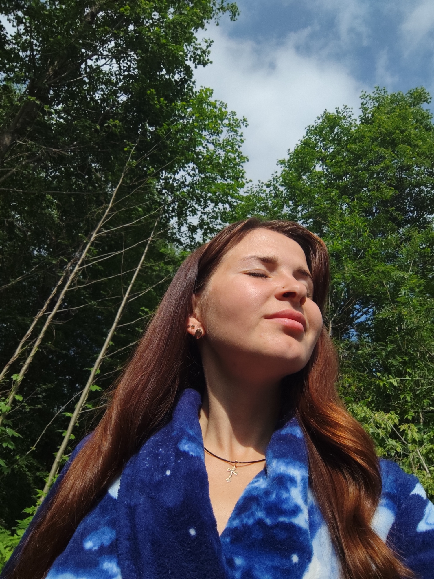 A young woman with long, reddish-brown hair is outdoors, with her eyes closed and face tilted upwards as if enjoying the sunlight. She is wearing a blue and white tie-dye robe and a gold cross necklace. The background consists of lush green trees and a blue sky with scattered white clouds. The location appears to be a natural, wooded area.
