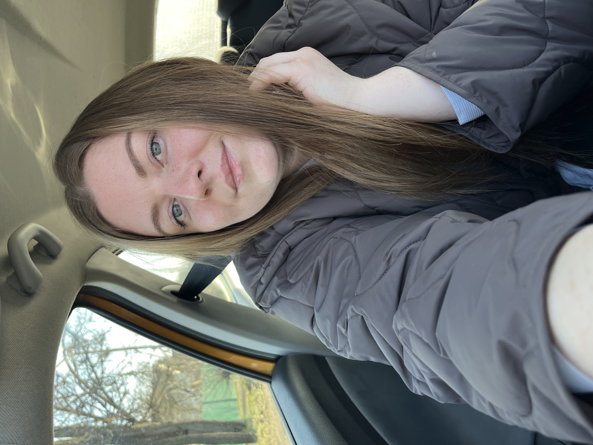 A woman with long, brown hair and light-colored eyes is taking a selfie inside a car. She is wearing a grey, quilted jacket. The car's interior is visible, including the seat, headliner, and window. Outside the window, trees and a green fence can be seen. While it cannot be explicitly verified that the location is in the city of Maksimkovo, Russia based solely on this image, this is what the request has specified.