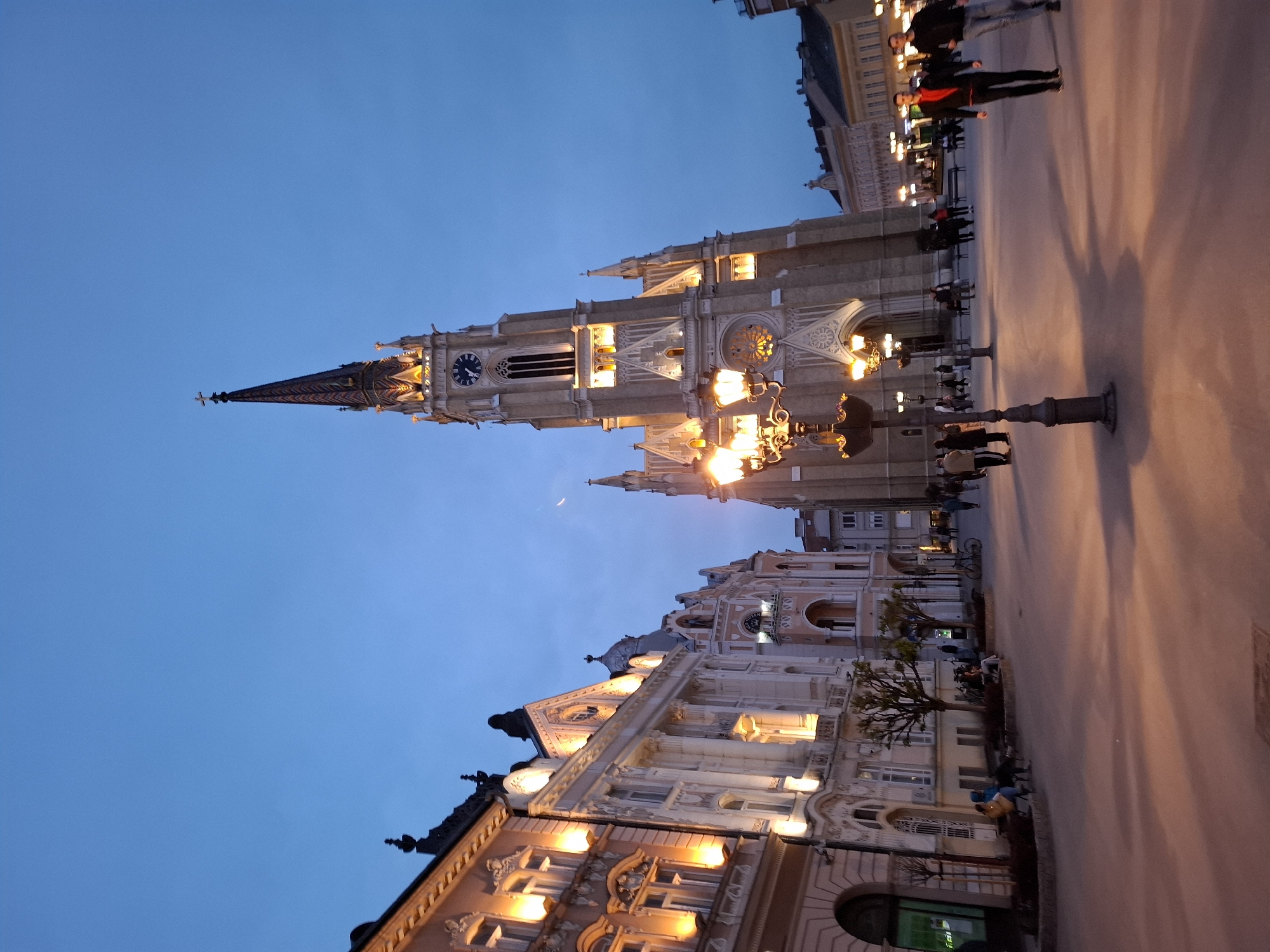 The image shows an evening view of buildings in Novi Sad, Serbia, with illuminated architectural details against a darkening blue sky. The prominent structure is the Name of Mary Church, featuring a tall spire with a clock and cross at the top. The foreground shows a lit building with ornate windows and architectural trim. The street level features a pavement area with a few people walking, as well as a lamp post. Buildings extend along the right of the photo with people standing around. A crescent moon is visible in the sky.