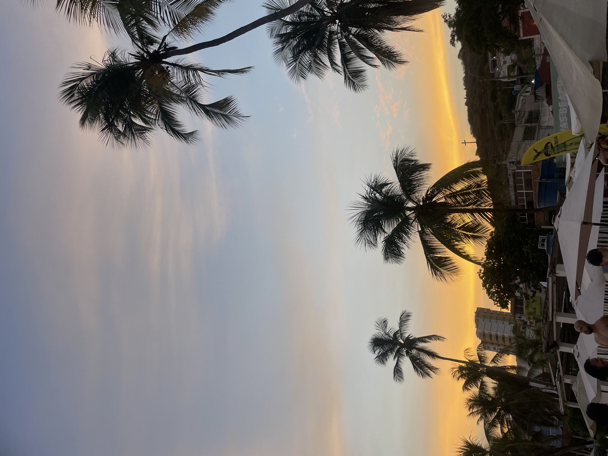 A twilight scene at a beach in Pampatar, Venezuela, features a vibrant sky transitioning from bright orange at the horizon to soft pinks and blues overhead. Several palm trees, silhouetted against the colorful sky, are scattered throughout the frame. In the background, buildings and a hillside with structures and a cross are visible. In the foreground, beach umbrellas and a few people are present, suggesting a coastal establishment. The image captures a serene moment at dusk.
