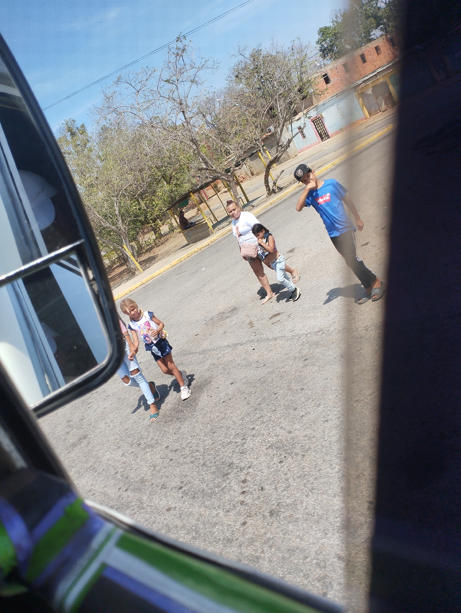 From the perspective of someone inside a vehicle, the photograph captures a street scene in Las Guevaras, Venezuela. Four people are walking on the road; a woman with a purse, a younger girl, a small child, and a boy wearing a black baseball cap and a blue shirt with 'Levis' written on it. The building in the background is light blue and has some brick details. Trees with bare branches are along the roadside. A structure appears to be present on the roadside. The vehicle's side mirror is visible in the left side of the frame. A portion of the interior is shown.