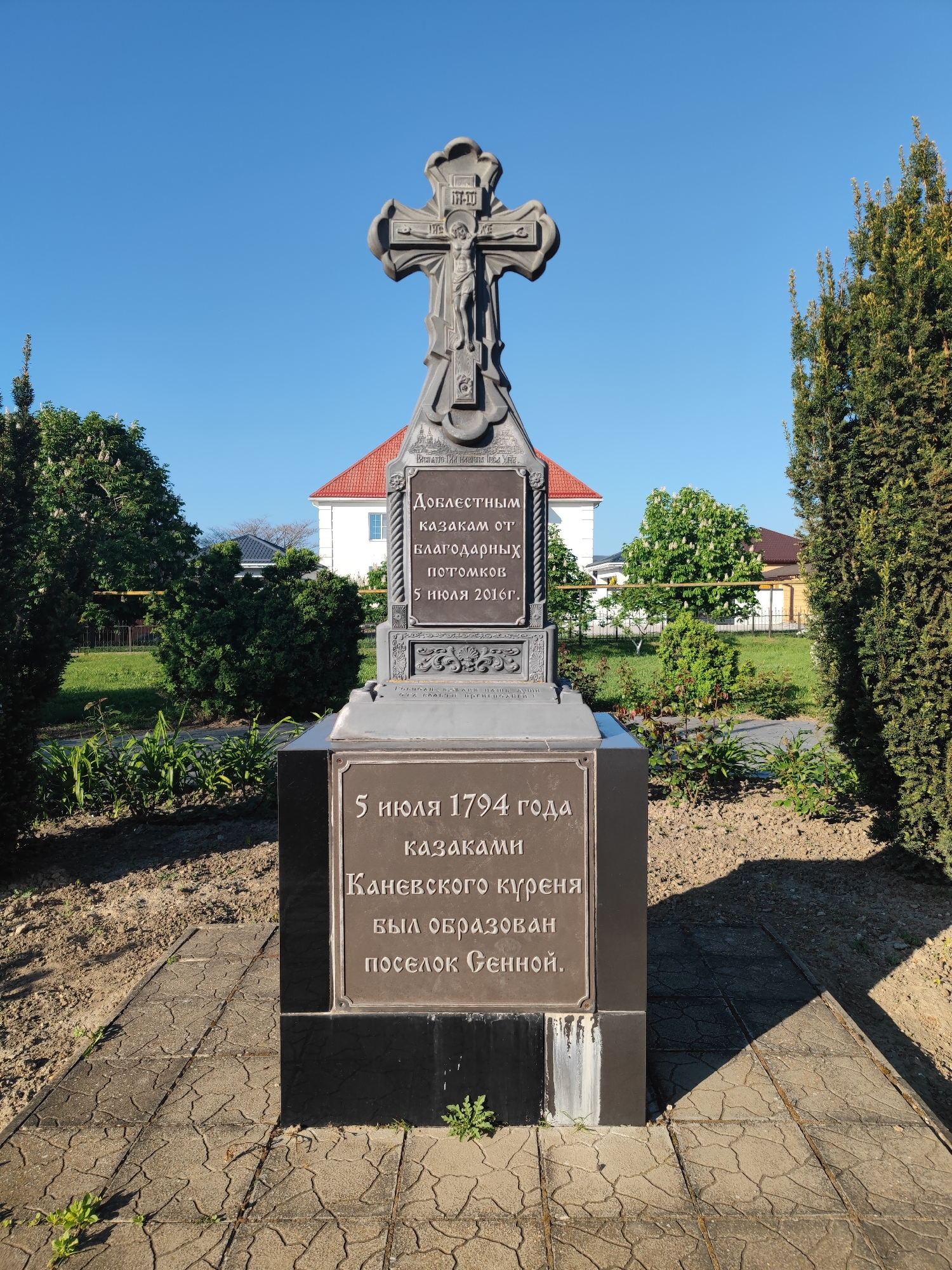 The image shows a monument in the city of Sennoy, Russia. The monument is dedicated to the Cossacks. It is made of dark metal and stone, with a cross on top. The monument has inscriptions in Russian, stating that the Cossacks of the Kanevskoy district founded the settlement of Sennoy on July 5, 1794. The monument was erected by grateful descendants on July 5, 2016. The monument is surrounded by greenery and trees. A house with a red roof can be seen in the background.