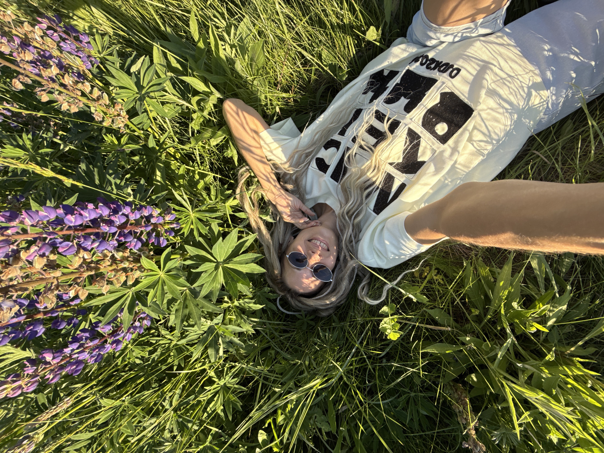An outdoor scene in Заволжье, Russia, depicts a fair-skinned female lying on her back in a field of tall green grass and purple lupine flowers. The image is a high-angle selfie, with one arm extending from the bottom of the frame, holding the camera. She has long, light-colored hair, possibly blonde dreadlocks or thick braids, spread around her head. She wears dark, round sunglasses and smiles directly at the camera. Her attire includes a light-colored (off-white or light green) short-sleeved t-shirt with a prominent black, multi-block graphic design on the front, including part of a word "D.D.R.ZO" vertically on the right. Light blue shorts are partially visible. The surrounding vegetation is dense, composed of vibrant green grass and numerous stalks of purple lupines, some in full bloom and others fading. Strong, direct sunlight illuminates the scene, casting distinct shadows from the foliage onto her face, arm, and shirt.