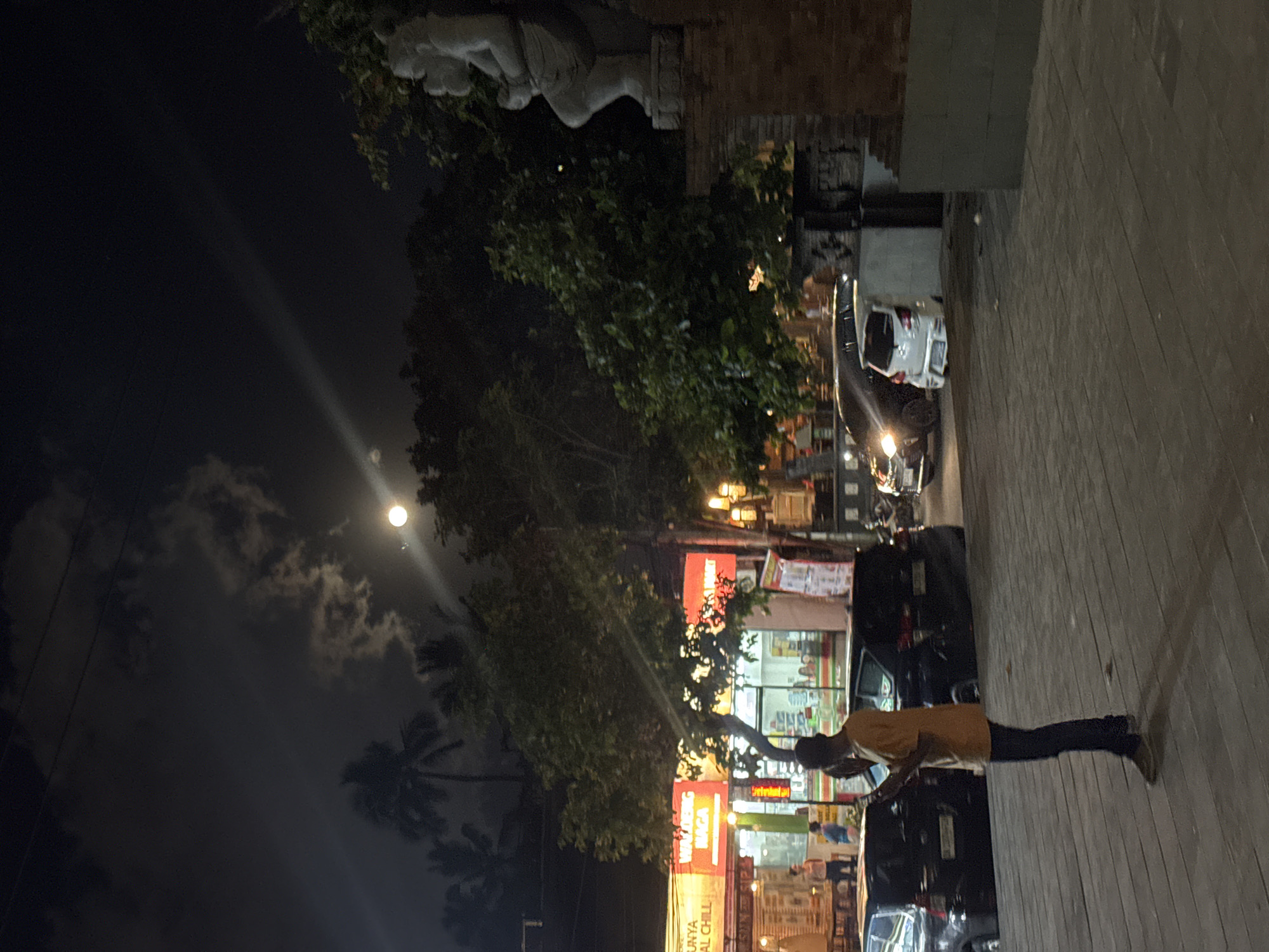 The image depicts a nighttime scene in Kuta, Indonesia, illuminated by streetlights and a bright full moon. A person, dressed in an orange shirt and dark pants, stands on a wooden boardwalk, looking at their phone. The boardwalk runs parallel to a street lined with shops and parked cars. The shops are brightly lit, with some displaying signs with Indonesian text. One sign, in large red lettering, reads "WARUNG NAGA". Another sign, also in red, partially visible, says "CHILL". The sky is dark, with scattered clouds and the moon shining prominently. In the background, palm trees are visible, suggesting a tropical environment. The overall atmosphere is that of a typical evening in a bustling tourist area, with the sounds of activity from the shops and street implied by the lighting and presence of people and vehicles.
