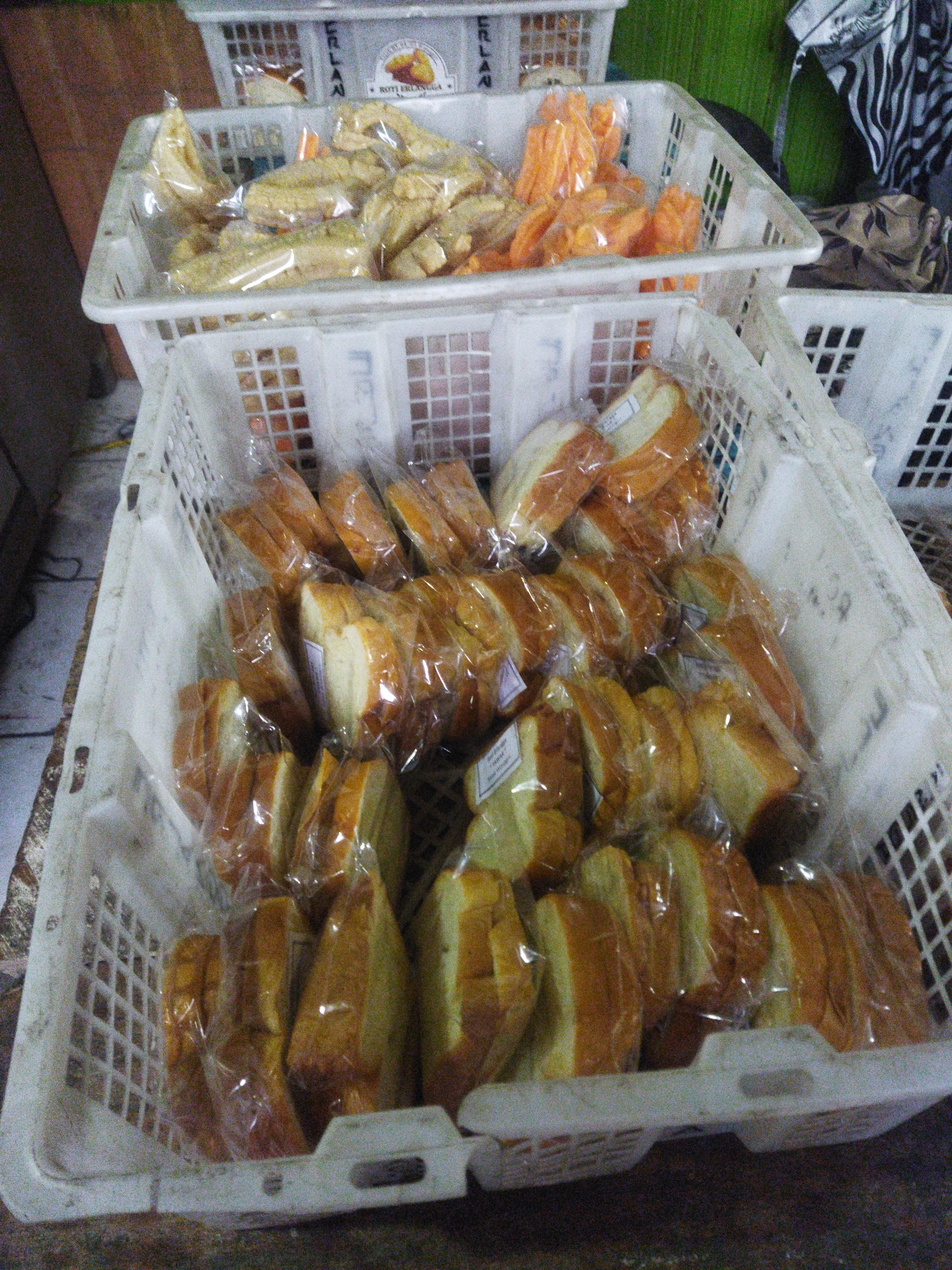 The image shows three white plastic baskets filled with packaged food items. The bottom basket is the largest and contains numerous slices of bread, each individually wrapped in clear plastic. Some of the bread slices have labels attached. The middle basket, slightly smaller, is filled with pieces of what appear to be dried snacks, possibly sweet potato or similar root vegetables, also in clear plastic packaging. The top basket, the smallest, is partially obscured but also contains similar dried snack items, some of which are orange in color. The baskets are stacked and appear to be arranged on a surface, possibly a market stall or countertop, given the slightly worn appearance of the surface in the foreground and the textured background. The overall impression is a display of food products for sale. The presence of the text "ROTI ERLANGA" on a label in the middle basket suggests the products might be from a bakery or food vendor with that name. Without further contextual information or visible landmarks, determining the precise location within the city of Wonogiri, Indonesia, is not possible from the image alone. The lighting suggests an indoor or shaded outdoor environment.