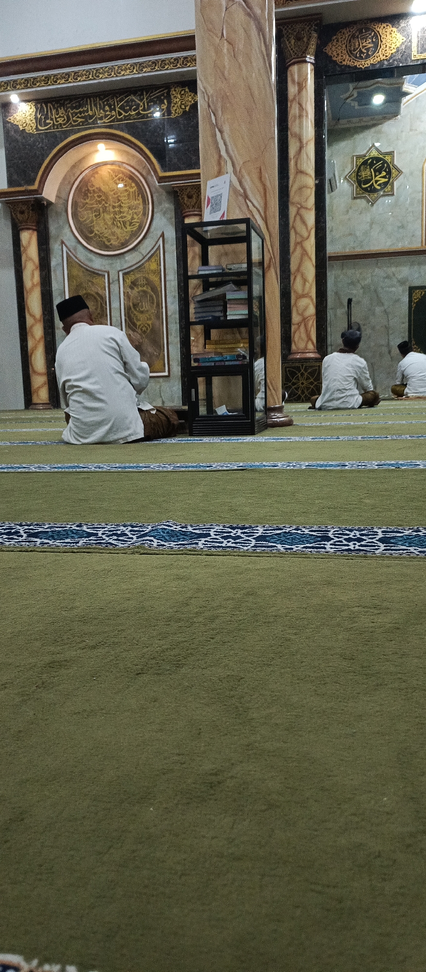 This image depicts the interior of a mosque, likely located in Arjasa, Indonesia. The foreground is dominated by a textured, olive-green carpet with a decorative blue and white striped pattern running horizontally across the frame. In the mid-ground, three individuals are seated on the carpet facing away from the camera. The leftmost individual is wearing a white thobe and a black peci (cap), facing towards what appears to be a prayer area with ornate wall decorations. To their right, two other individuals, also dressed in white and wearing dark caps, are seated on the carpet.
Behind the seated individuals, a tall, black, multi-tiered shelf unit stands prominently, filled with books and other items. The background features a wall with marble-patterned pillars and intricate Islamic calligraphy and geometric designs. A large, circular motif with gold calligraphy is visible on the wall behind the leftmost seated person. The lighting suggests an indoor environment, likely during daytime hours. The overall atmosphere is one of quiet contemplation and prayer.