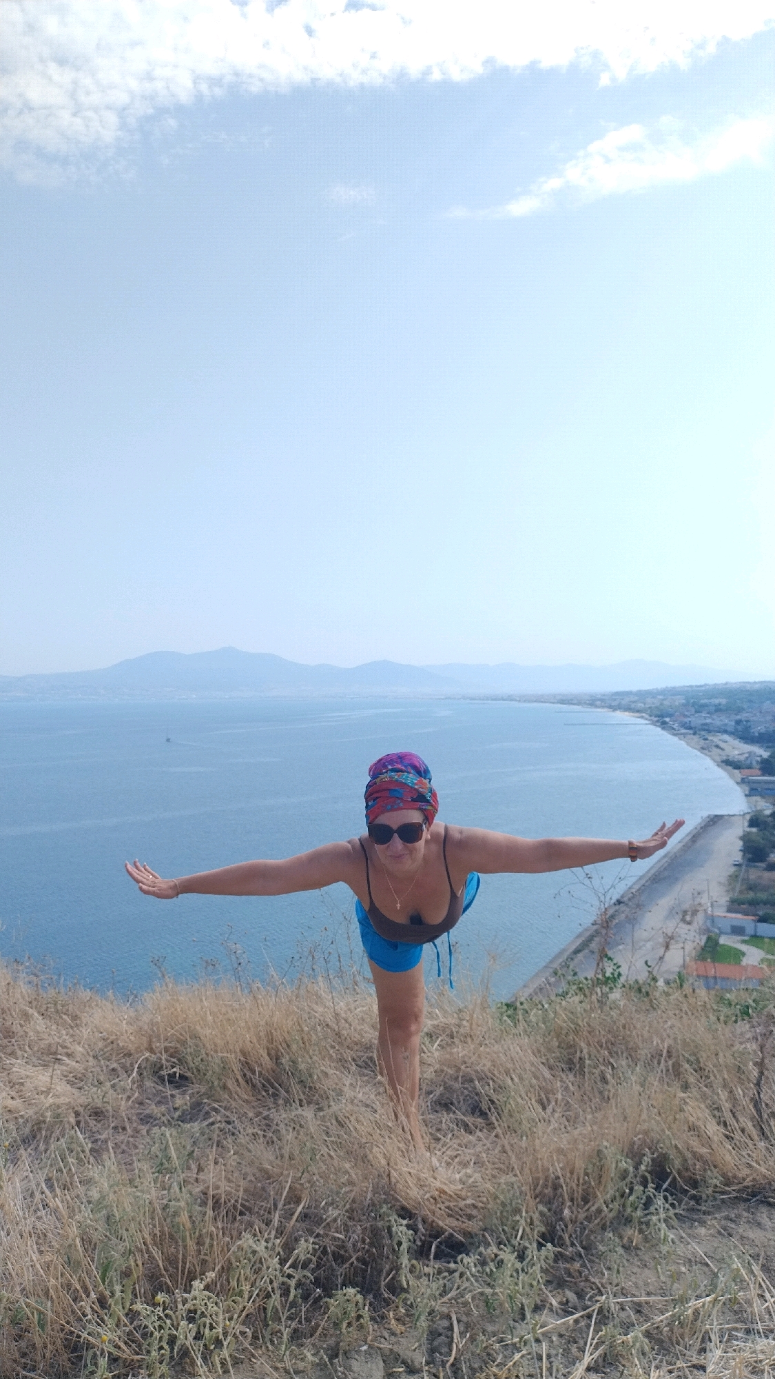 A woman stands on a grassy hill overlooking the Aegean Sea in Agia Triada, Greece, performing a yoga pose. She is wearing a brown tank top, blue shorts, sunglasses, and a colorful headscarf. Her arms are extended outwards for balance, and her body is angled slightly forward. The sea stretches out before her, meeting a distant coastline dotted with buildings and backed by hazy mountains. The sky is a pale blue with scattered clouds. The foreground is dominated by dry, golden grass.