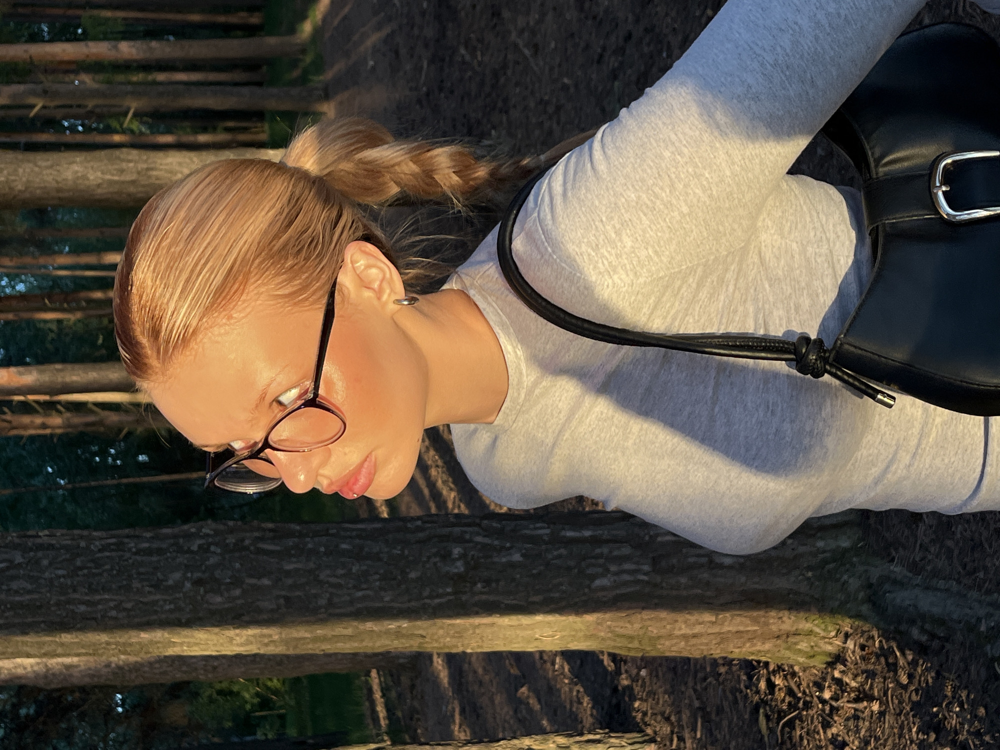 A fair-skinned woman with blonde hair pulled back in a ponytail is captured in an outdoor setting. She is wearing glasses and a light grey long-sleeved shirt. A black leather bag is slung over her shoulder. The environment appears to be wooded, with fallen logs and foliage visible. The location is Chaikovsky, Russia.